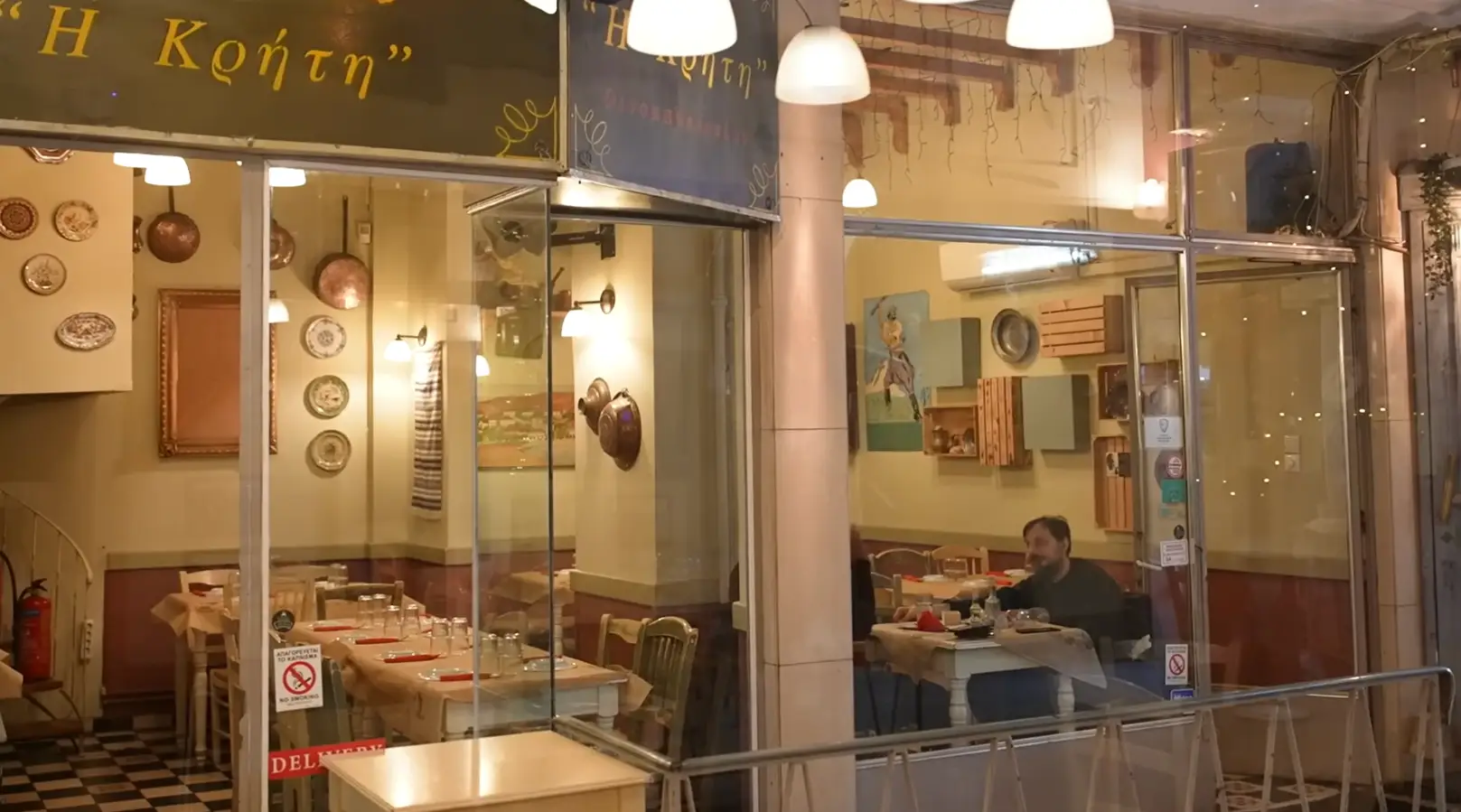 Old restuarant in Greece with plates and copper pots on the walls, a person sitting at a table dining on a black and white checkered floor.