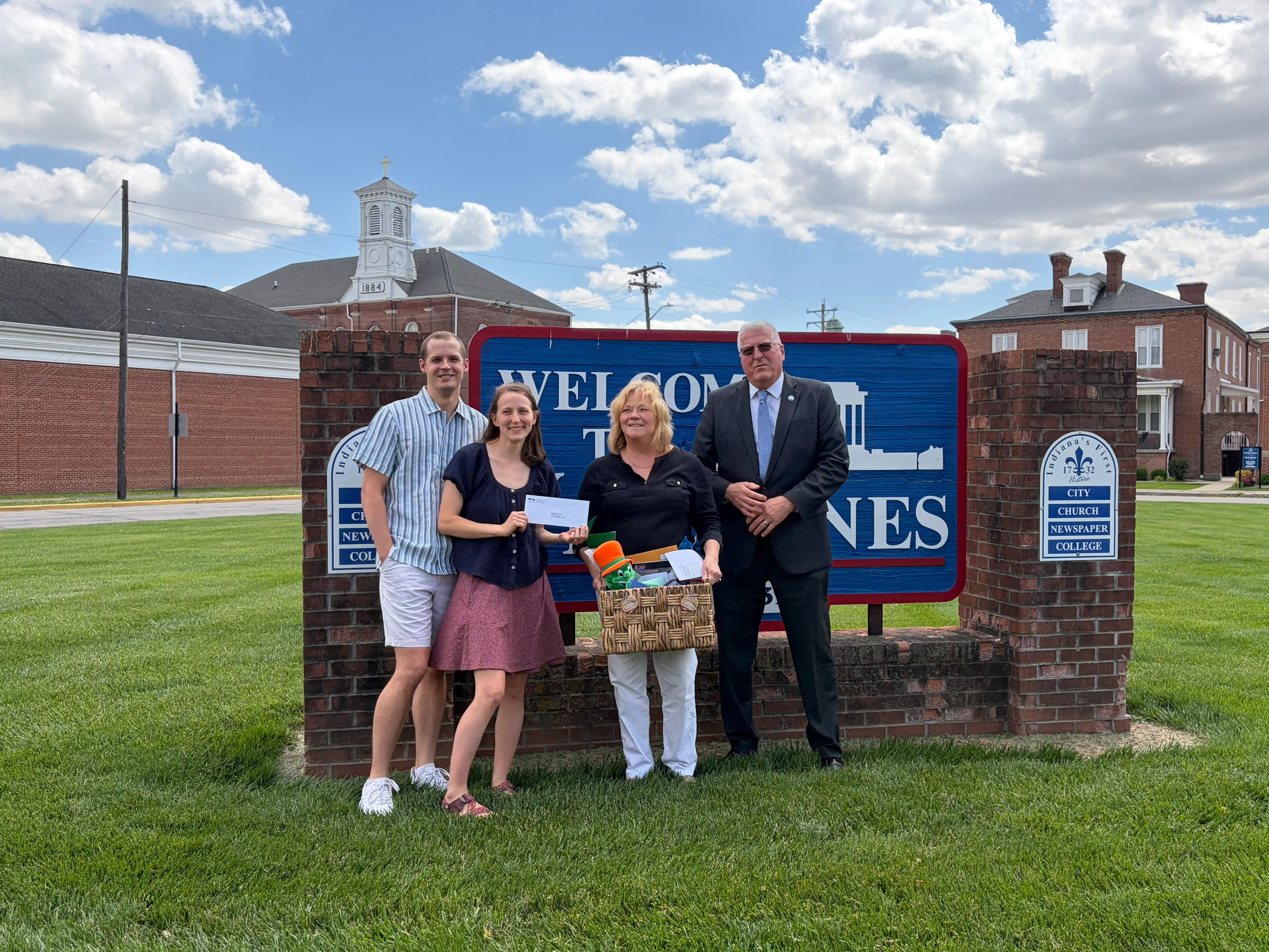 Four people pose on a lawn in front of a brick welcome sign, with an envelope and gift basket visible.
