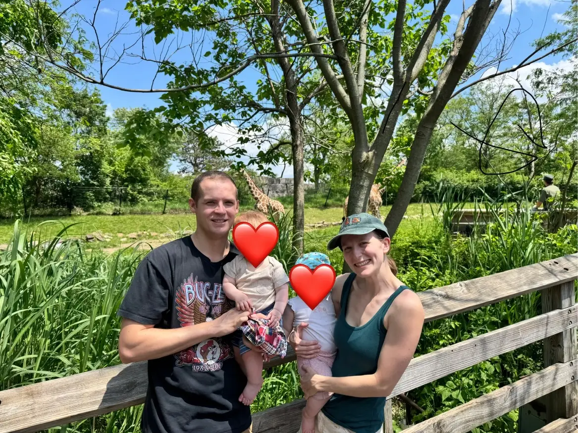 Two adults pose on a wooden walkway with two faces covered by heart stickers near giraffes and greenery.