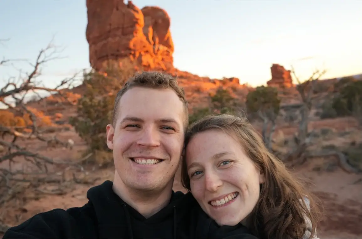 Two smiling people take a selfie in front of sunlit red sandstone formations in a desert landscape.