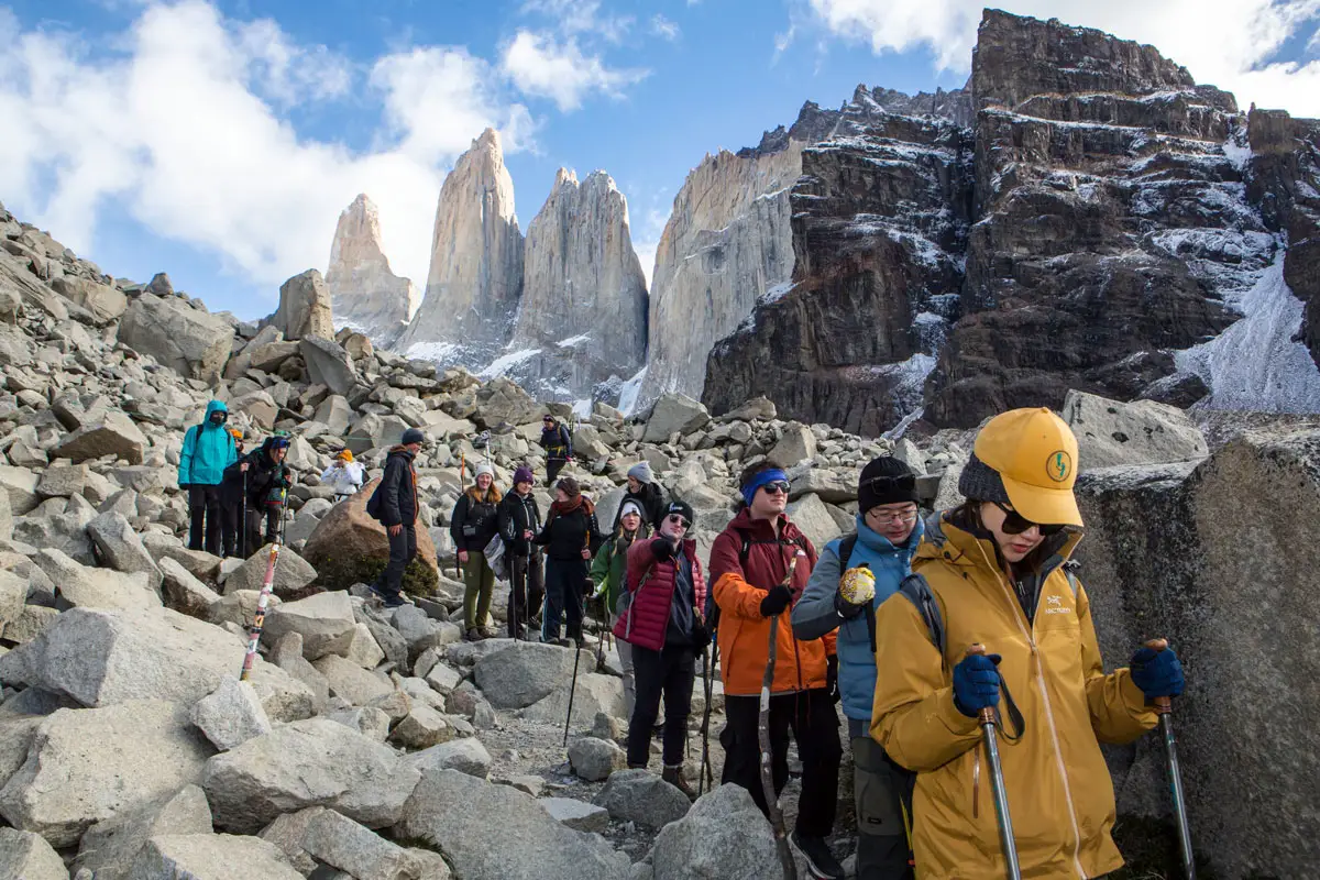 Hikers with trekking poles traverse a rocky trail beneath the Torres del Paine granite peaks.