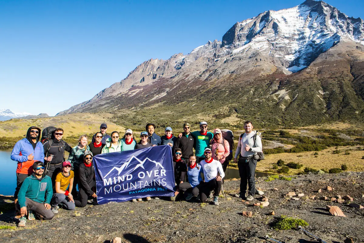 Group of hikers pose with a Mind Over Mountains banner beneath a snow-capped mountain.