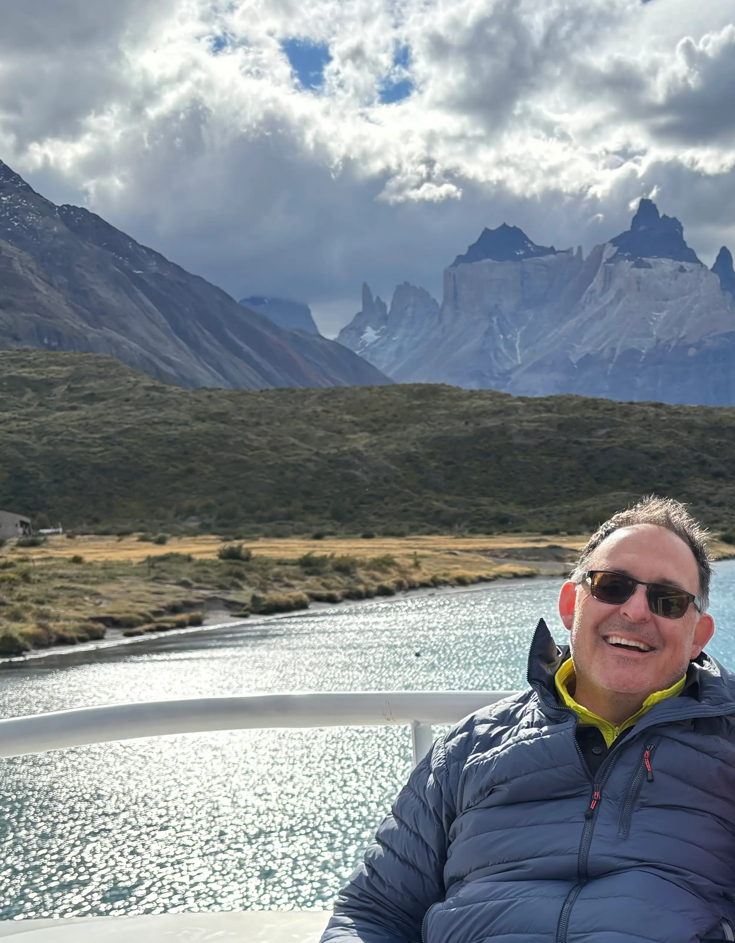 Ricardo Moya-Quiroga in sunglasses sits by a sparkling lake below dramatic Torres del Paine mountains.
