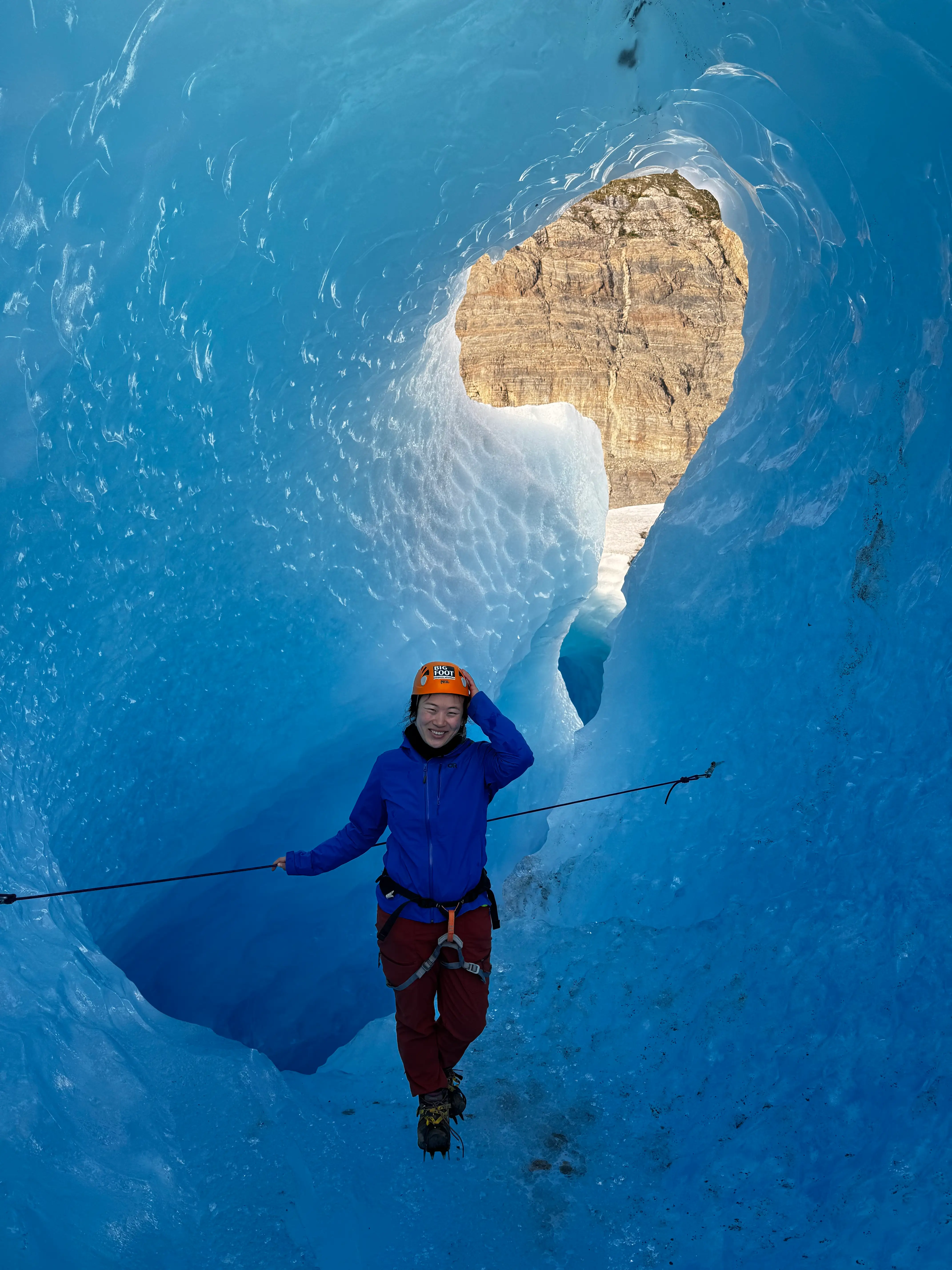 Ting Cui with helmet and rope standing inside a vivid blue glacier ice cave opening toward rocky cliffs.