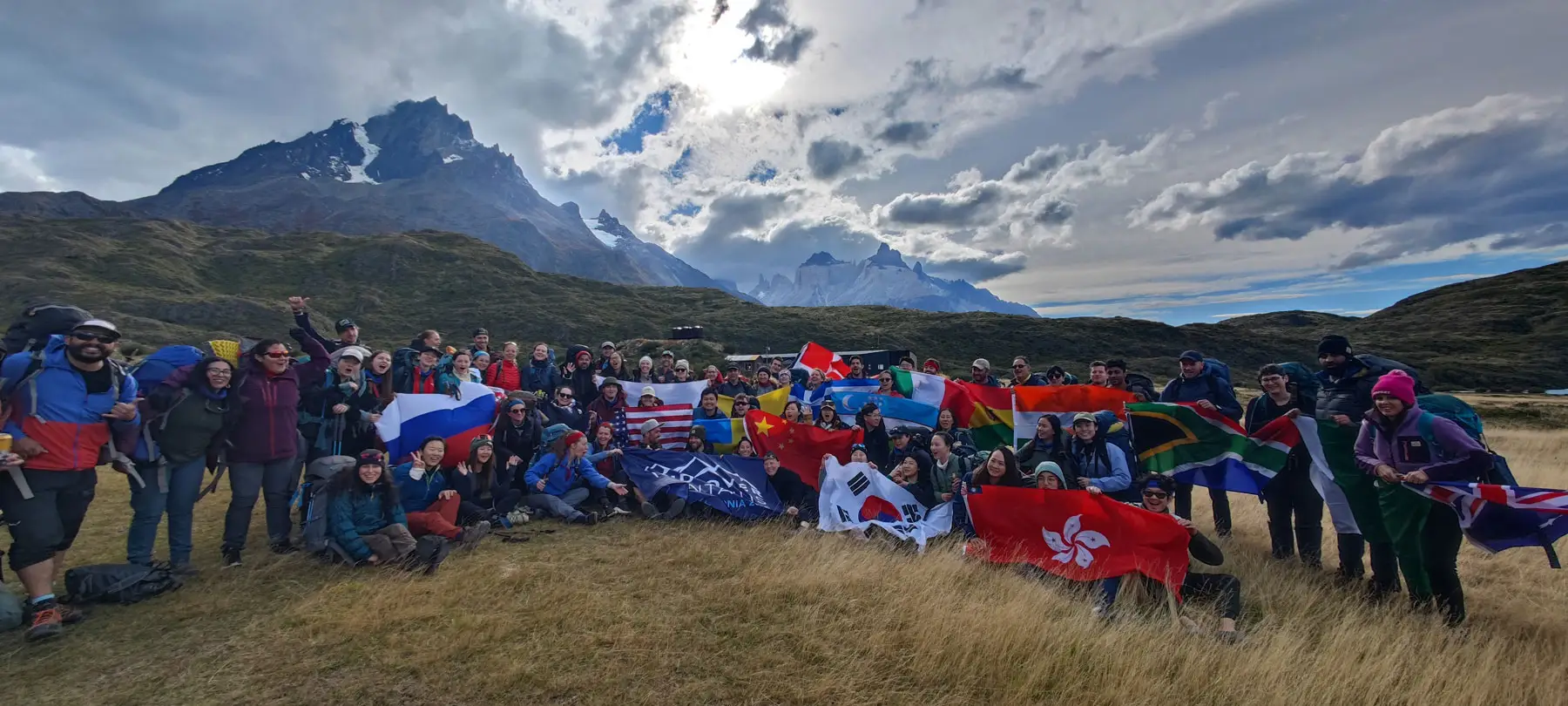 Large group of hikers poses with national flags in a grassy mountain landscape under a cloudy sky.