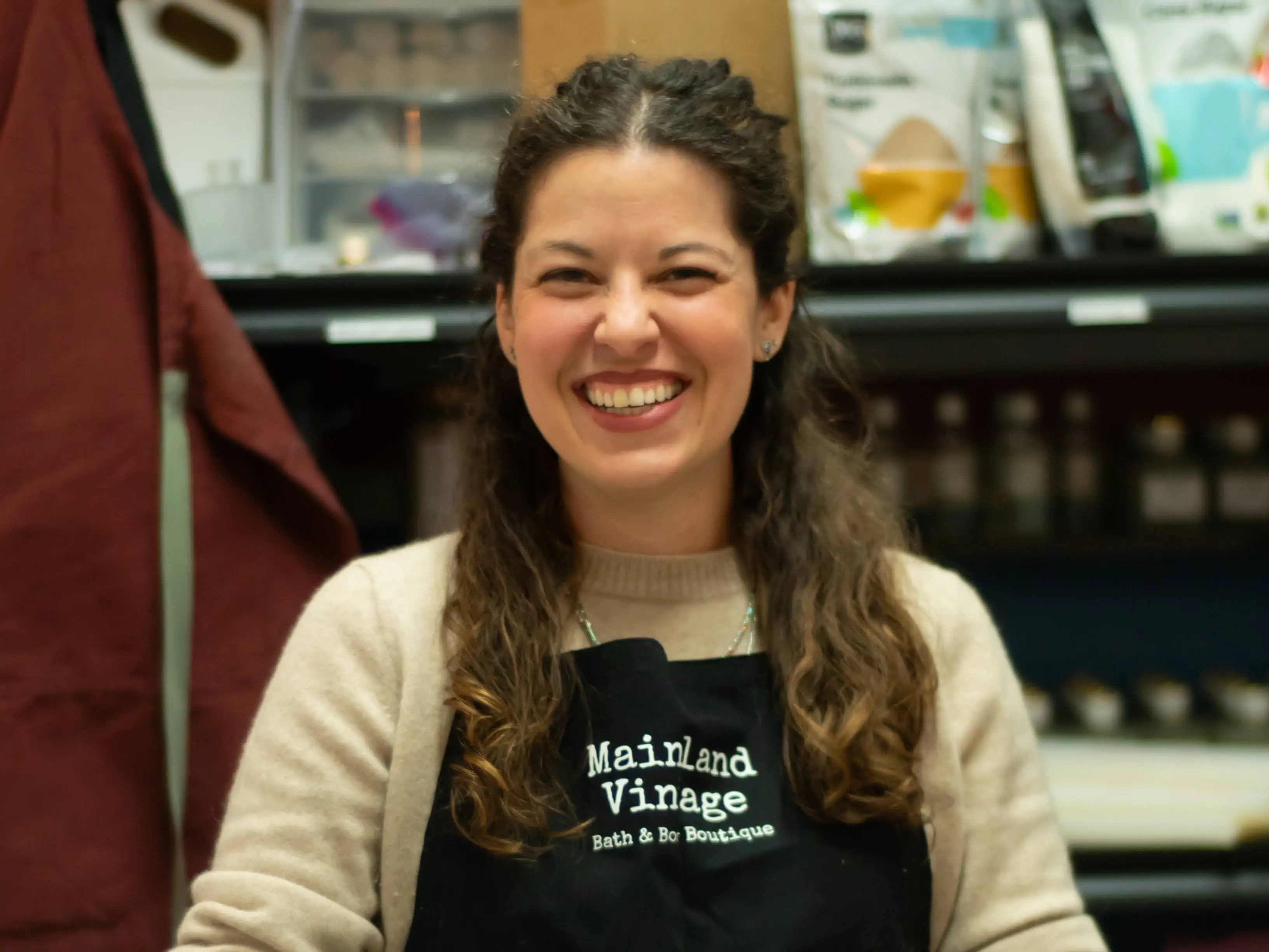 Smiling woman in a Mainland Vintage apron stands behind a counter with bottles and mixing supplies.