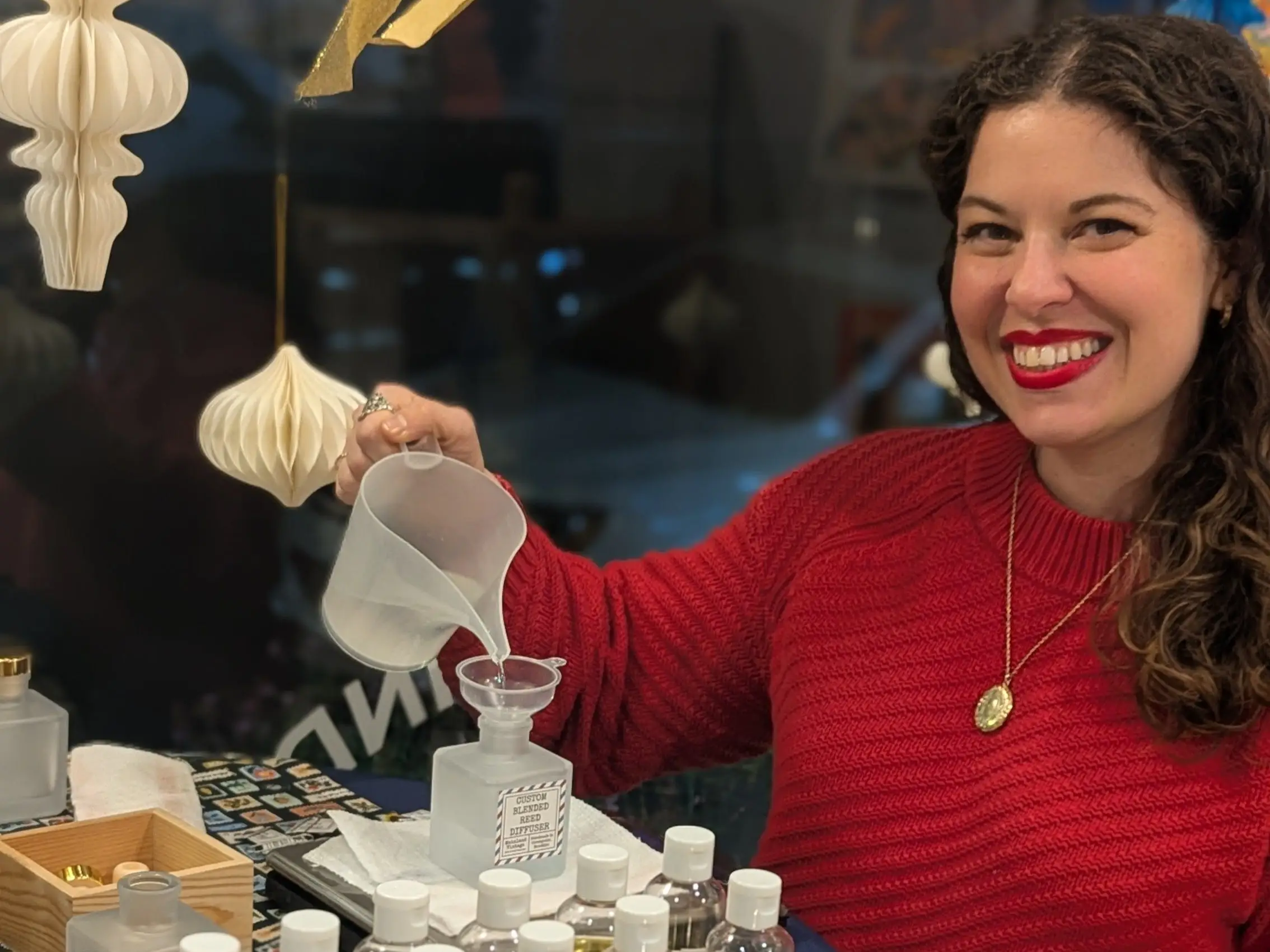 Smiling person in a red sweater pours liquid into a glass bottle at a table of fragrance bottles.