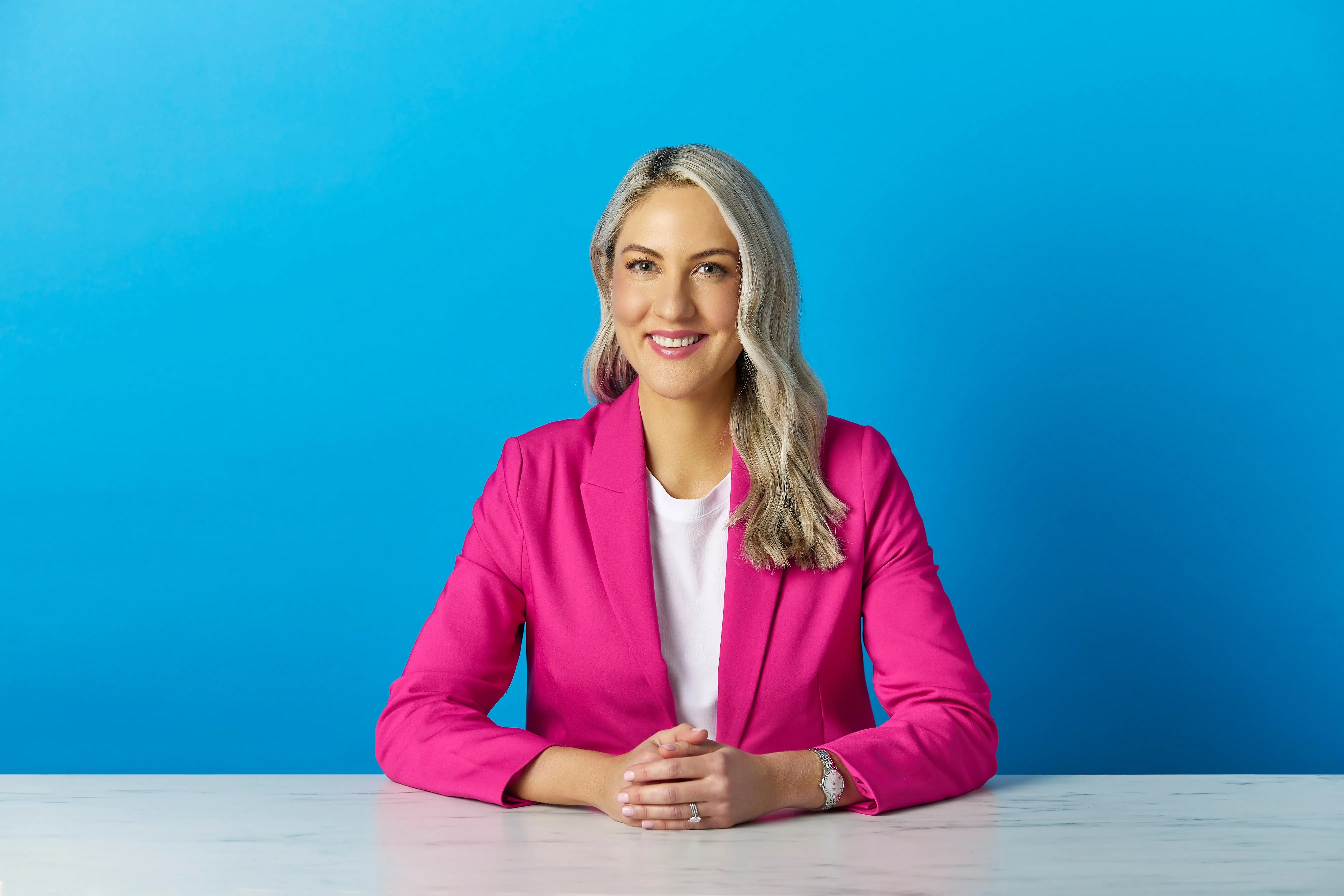 Smiling person in a bright pink blazer sits at a white table against a vivid blue studio backdrop.