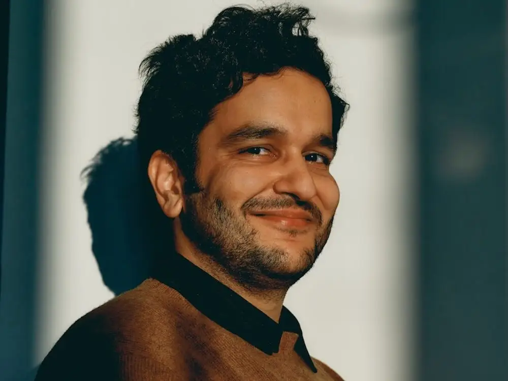 Smiling person in a brown sweater poses against a sunlit wall with strong shadow contrast.