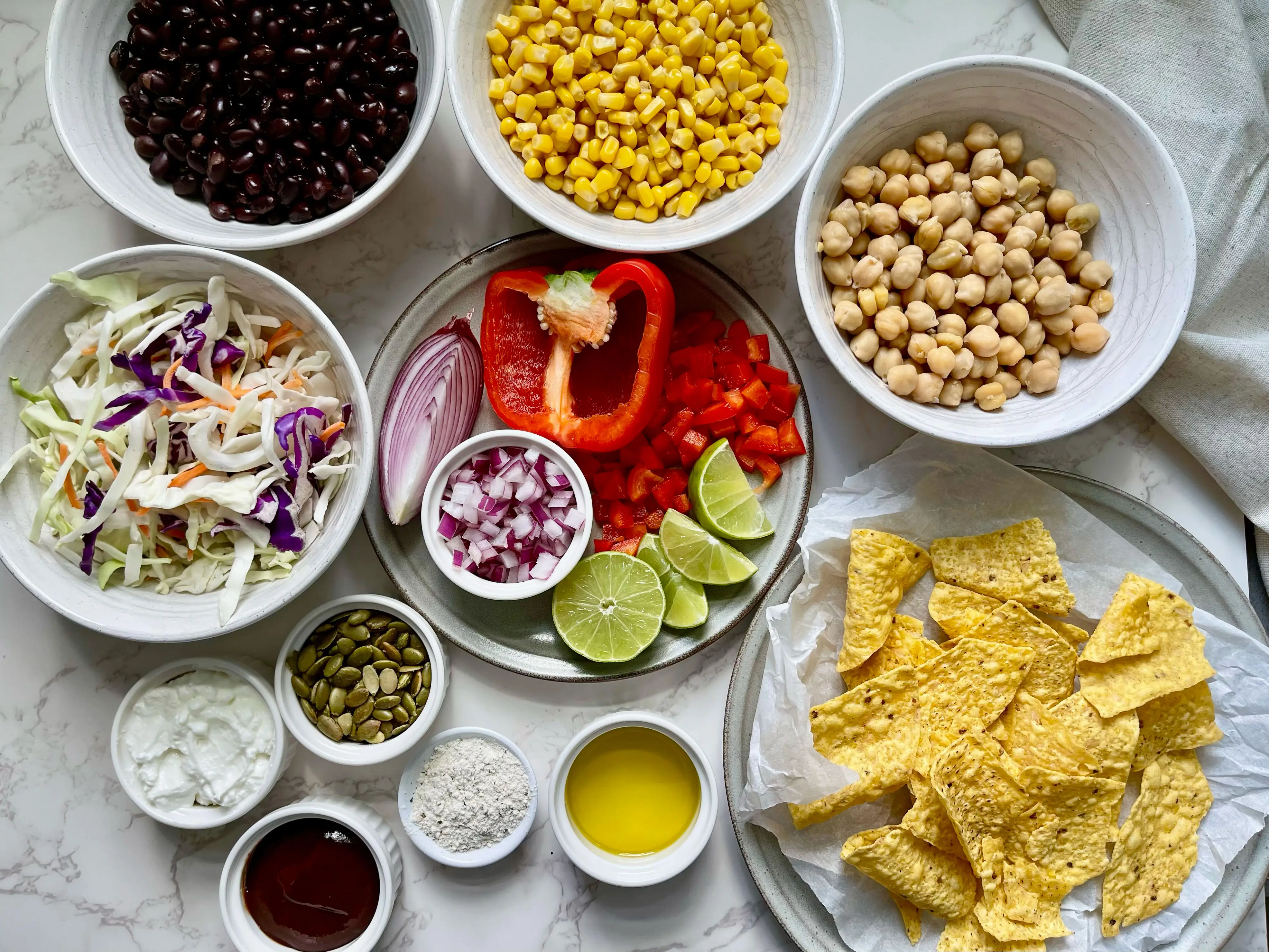 Bowls of black beans, corn, chickpeas, cabbage, red pepper, onion, lime, tortilla chips, and assorted spices and seeds arranged on a countertop.