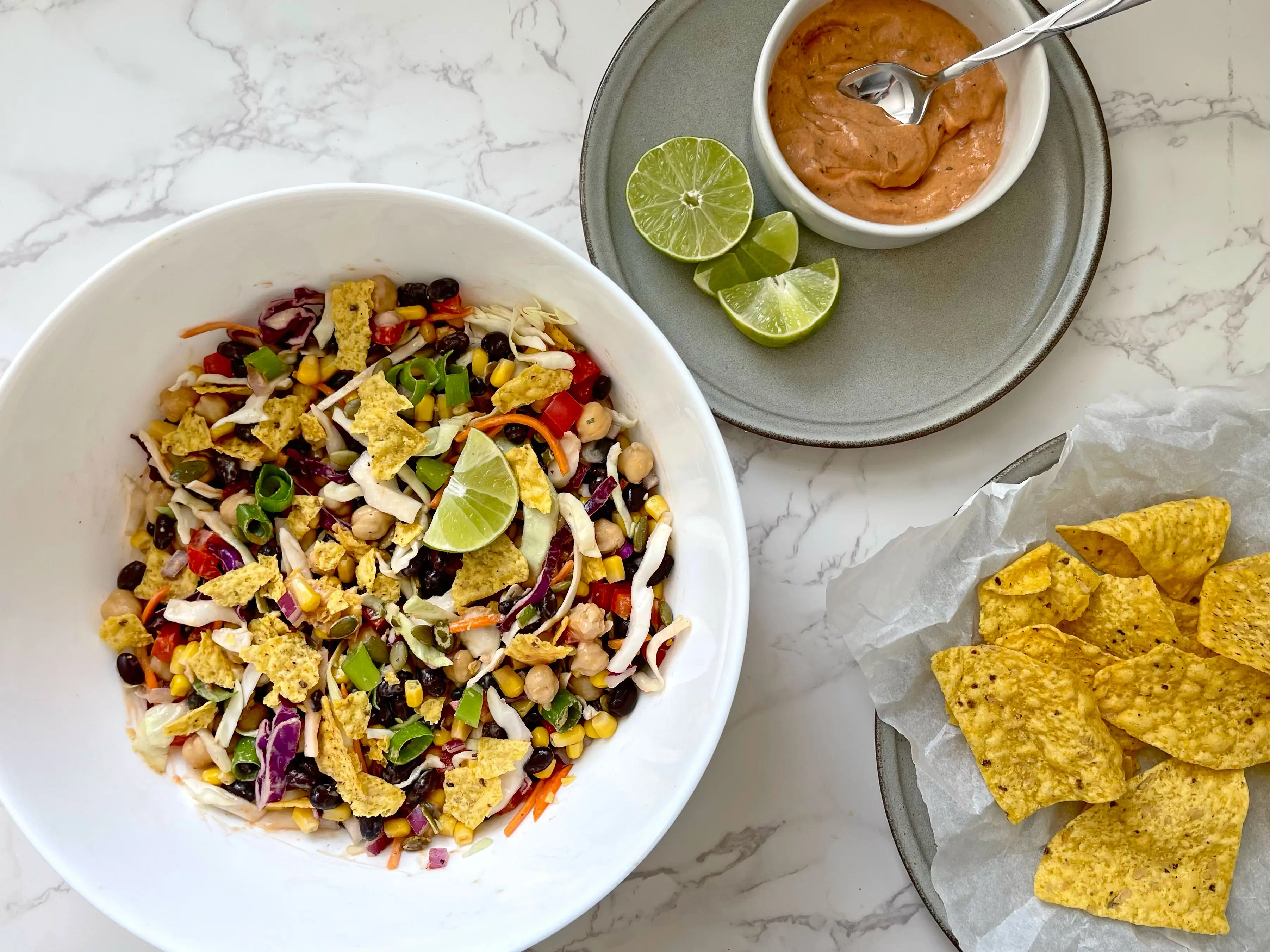 A bowl of bean salad, a ramekin of sauce, and a plate of tortilla chips on a countertop.