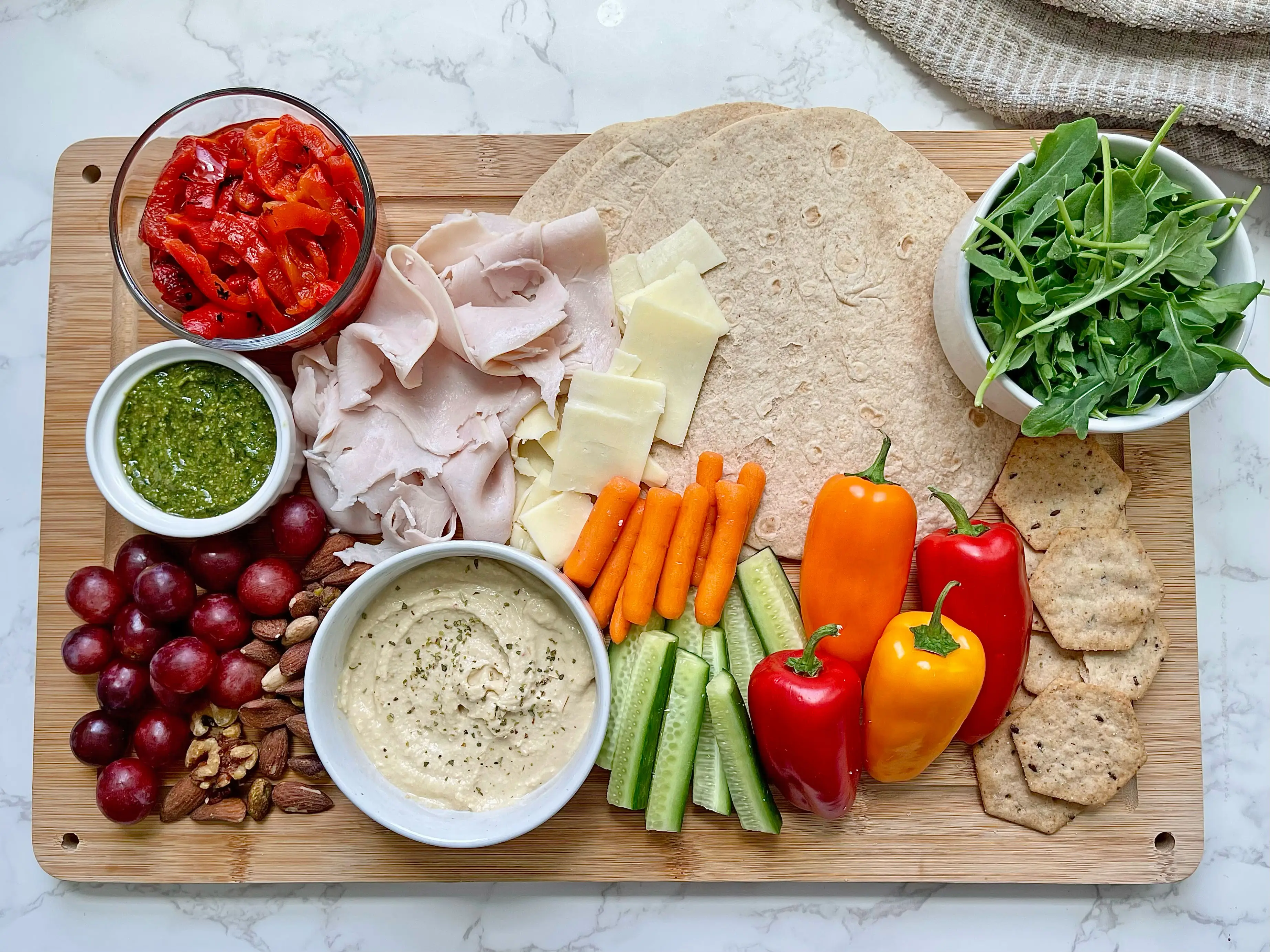 A cutting board with tortillas, sliced turkey, cheese, roasted peppers, pesto, hummus, grapes, nuts, carrots, sliced cucumber, arugula, and crackers.