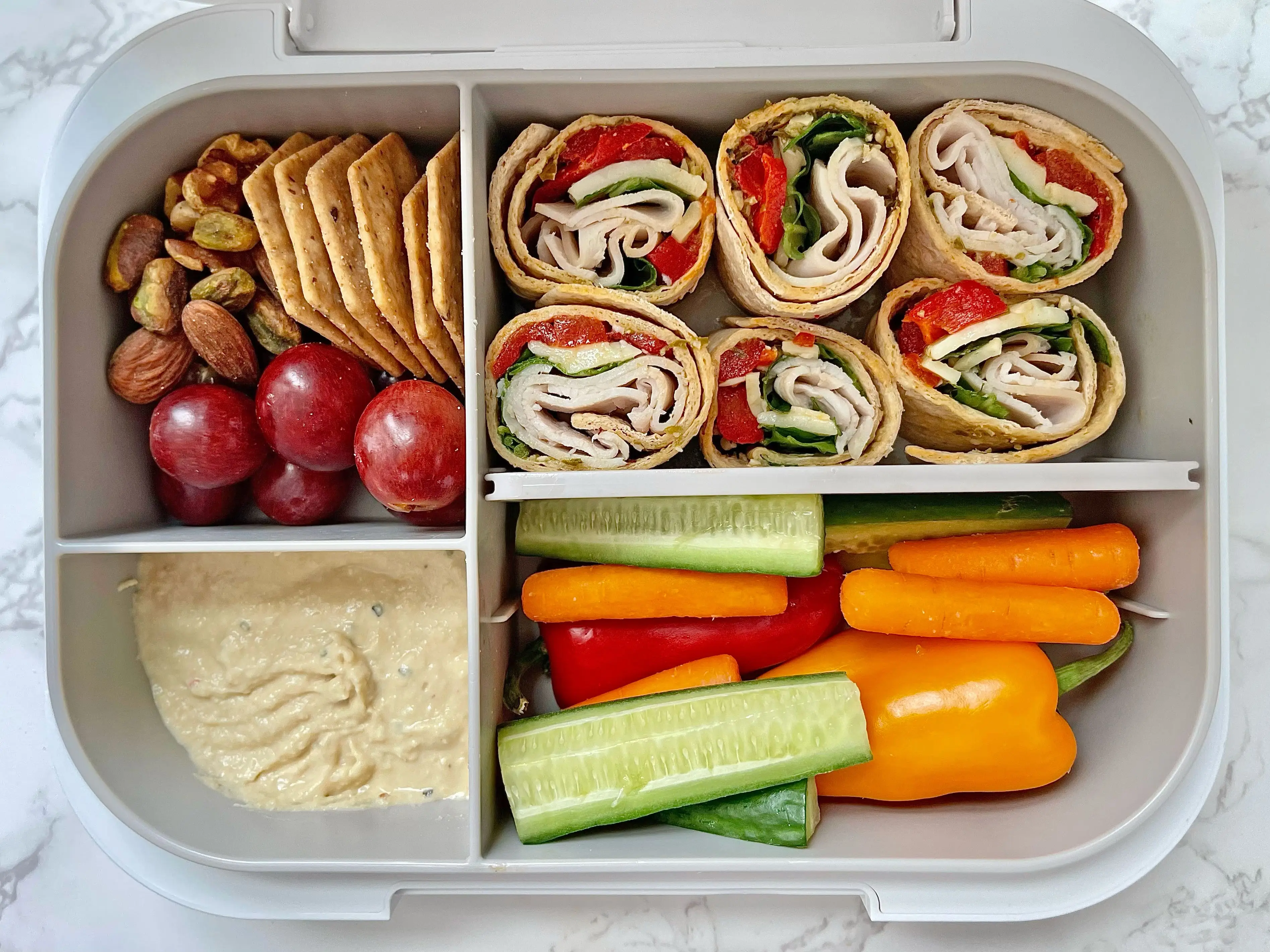 A bento box with a section of grapes, nuts, and crackers, a section of turkey-and-veggie pinwheels, a section of hummus, and a section of vegetables.