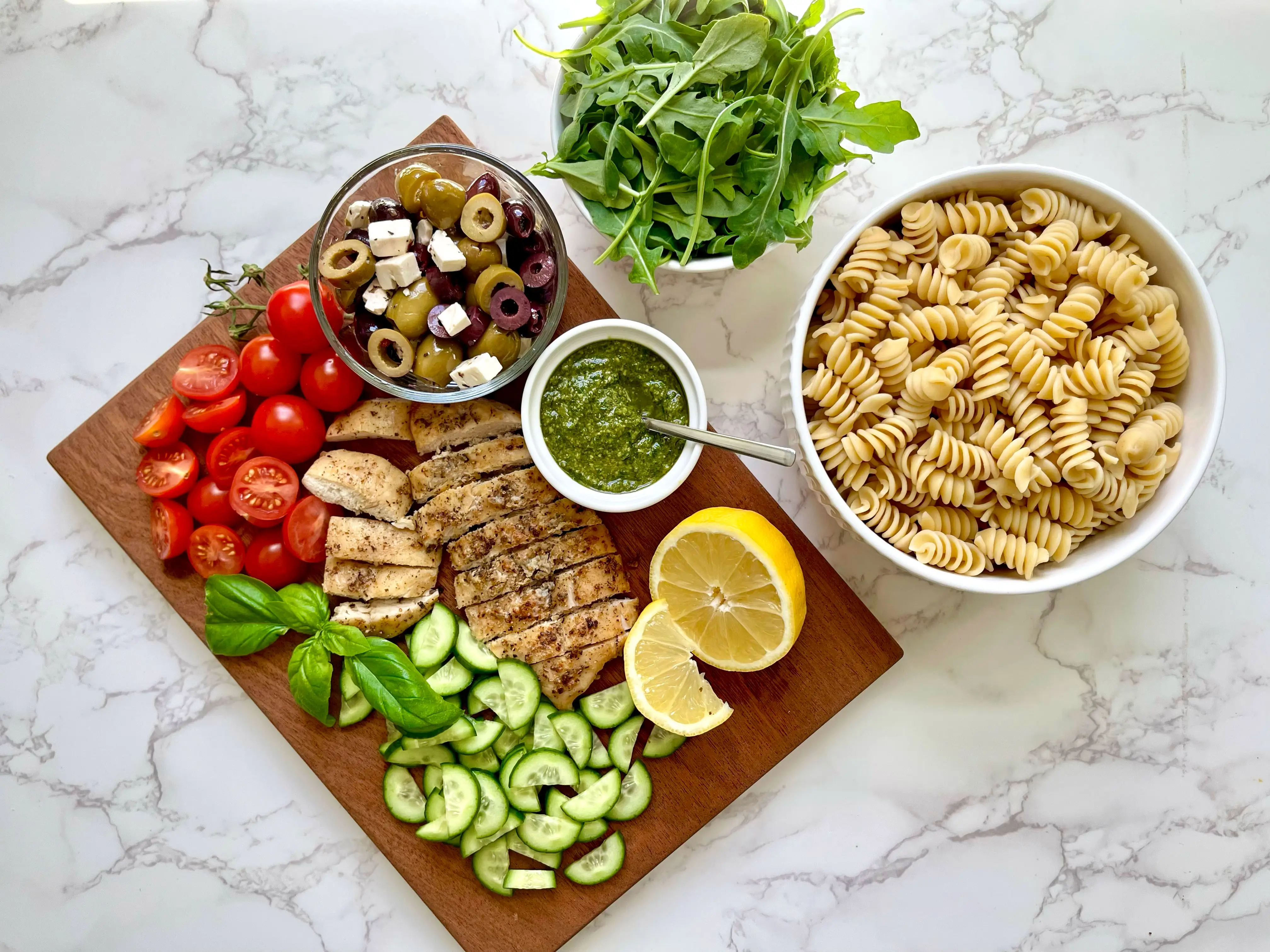Pasta salad ingredients with rotini, grilled chicken, tomatoes, cucumbers, olives, feta, arugula, pesto, basil, and lemon.