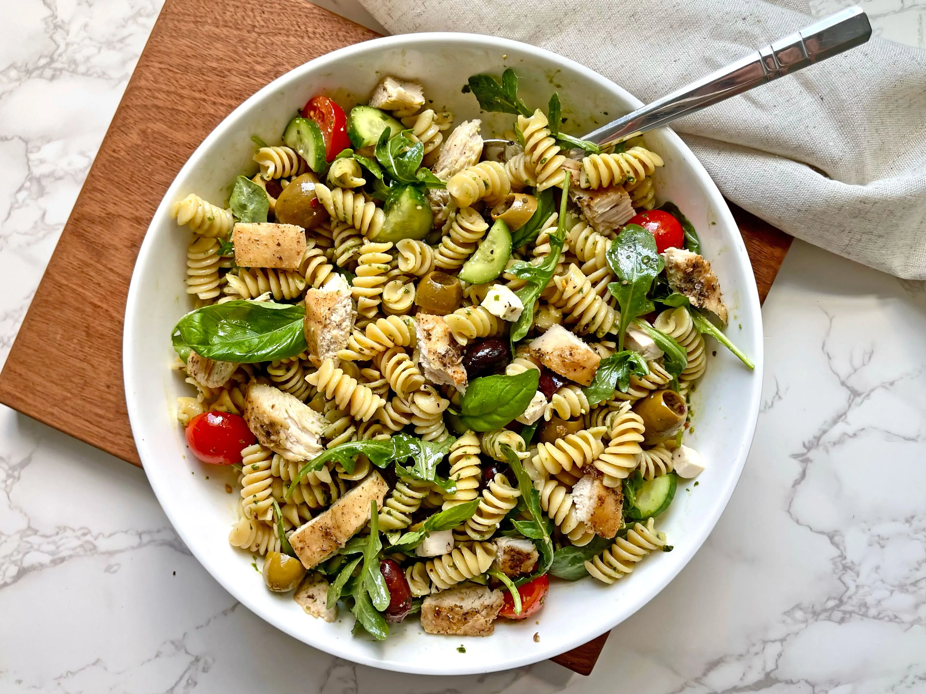 A bowl of pasta salad with chicken, arugula, feta, olives, and tomatoes.