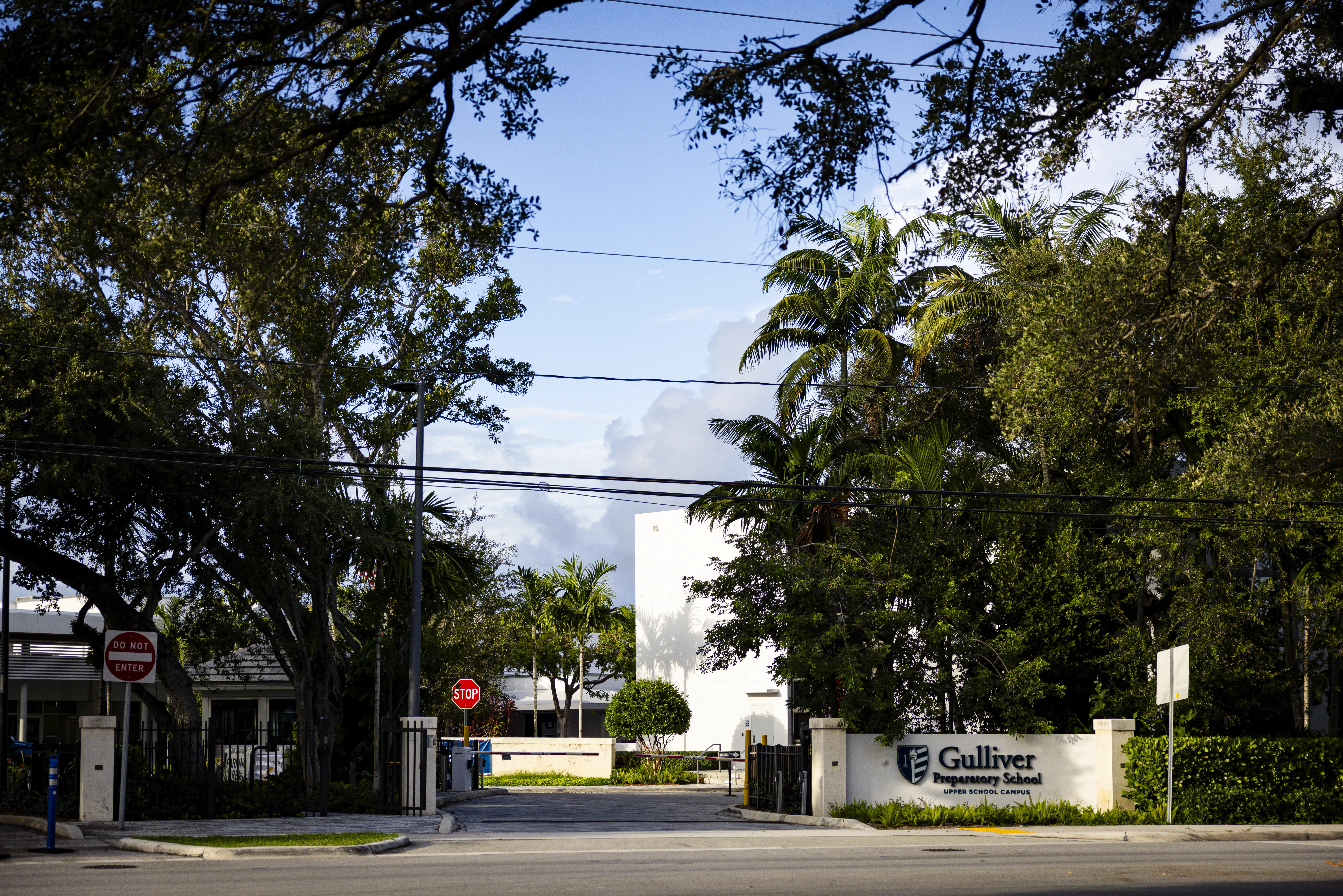 Tree-lined street entrance to Gulliver Preparatory School with campus sign, gates, palm trees, and stop signs.