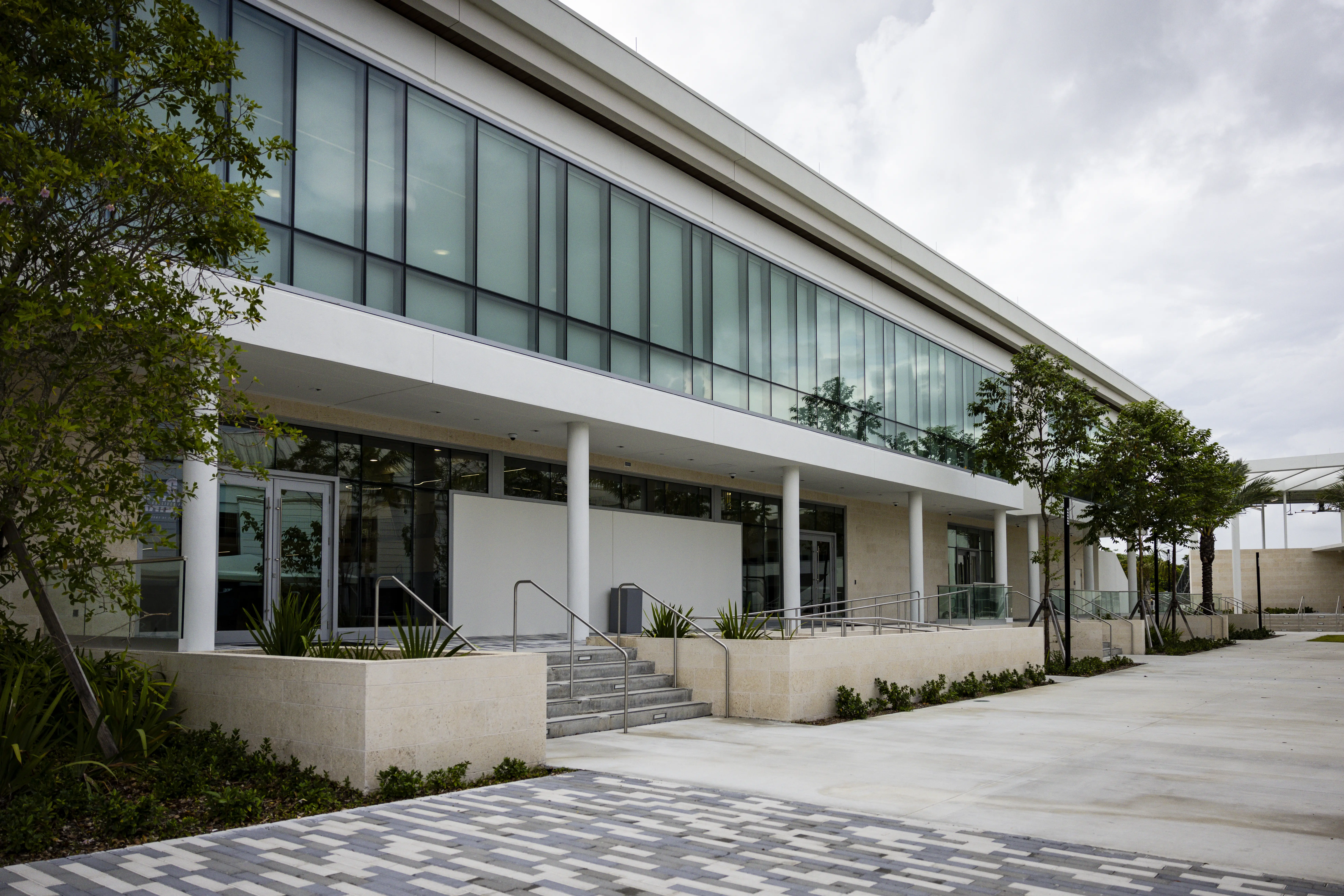 Modern two-story building with glass windows, white columns, stairs, and landscaped walkway under a cloudy sky.