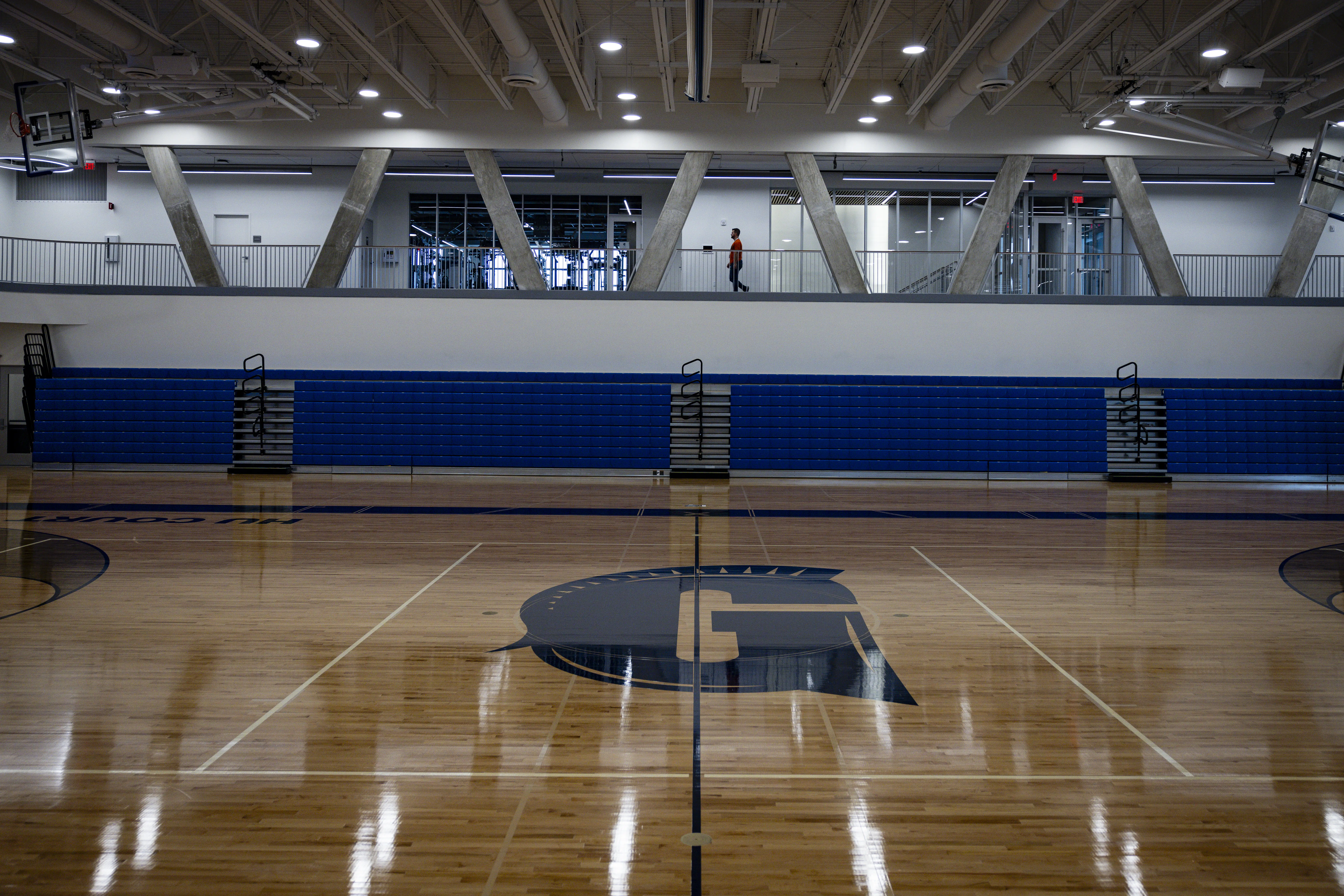 Empty indoor gymnasium with polished wood court, blue bleachers, and an upper-level walkway.