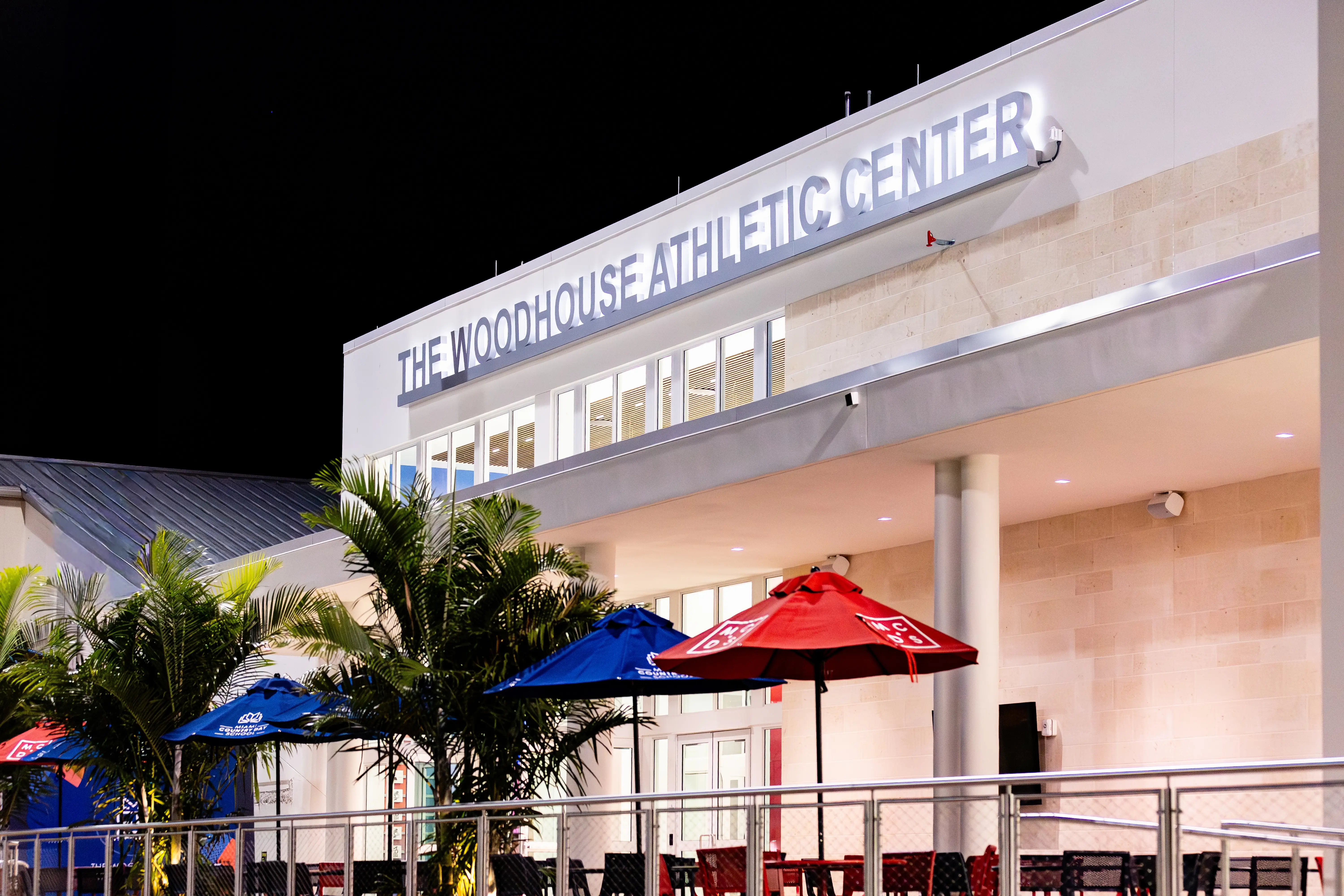 Nighttime exterior of The Woodhouse Athletic Center, Miami Country Day School, with patio umbrellas and palm landscaping near the entrance.