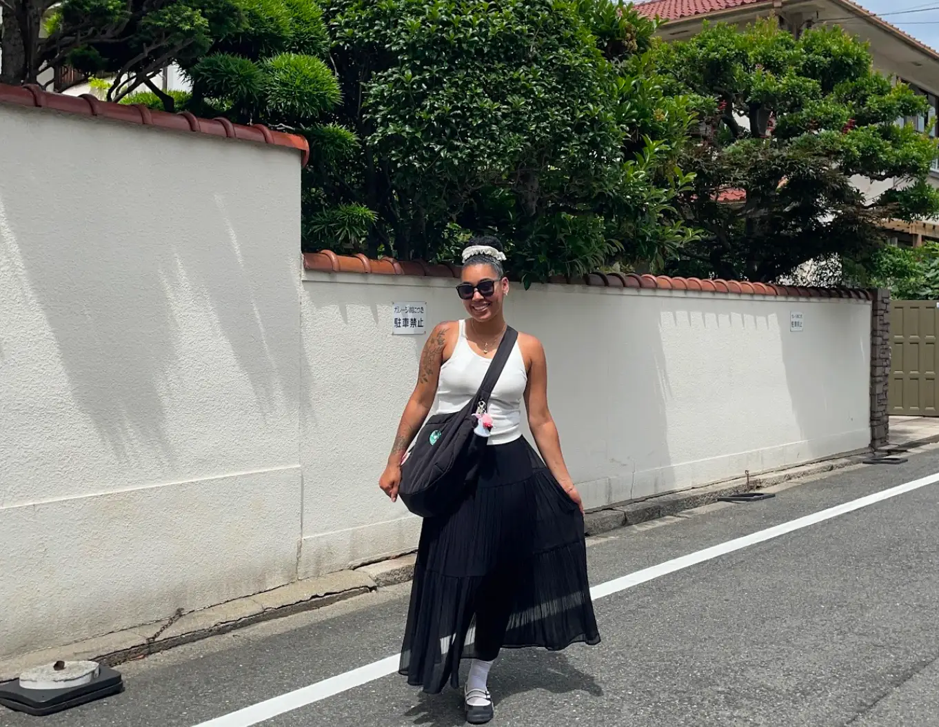 The writer wearing a black skirt and white top, and posing in front of a white wall and trees in Japan.