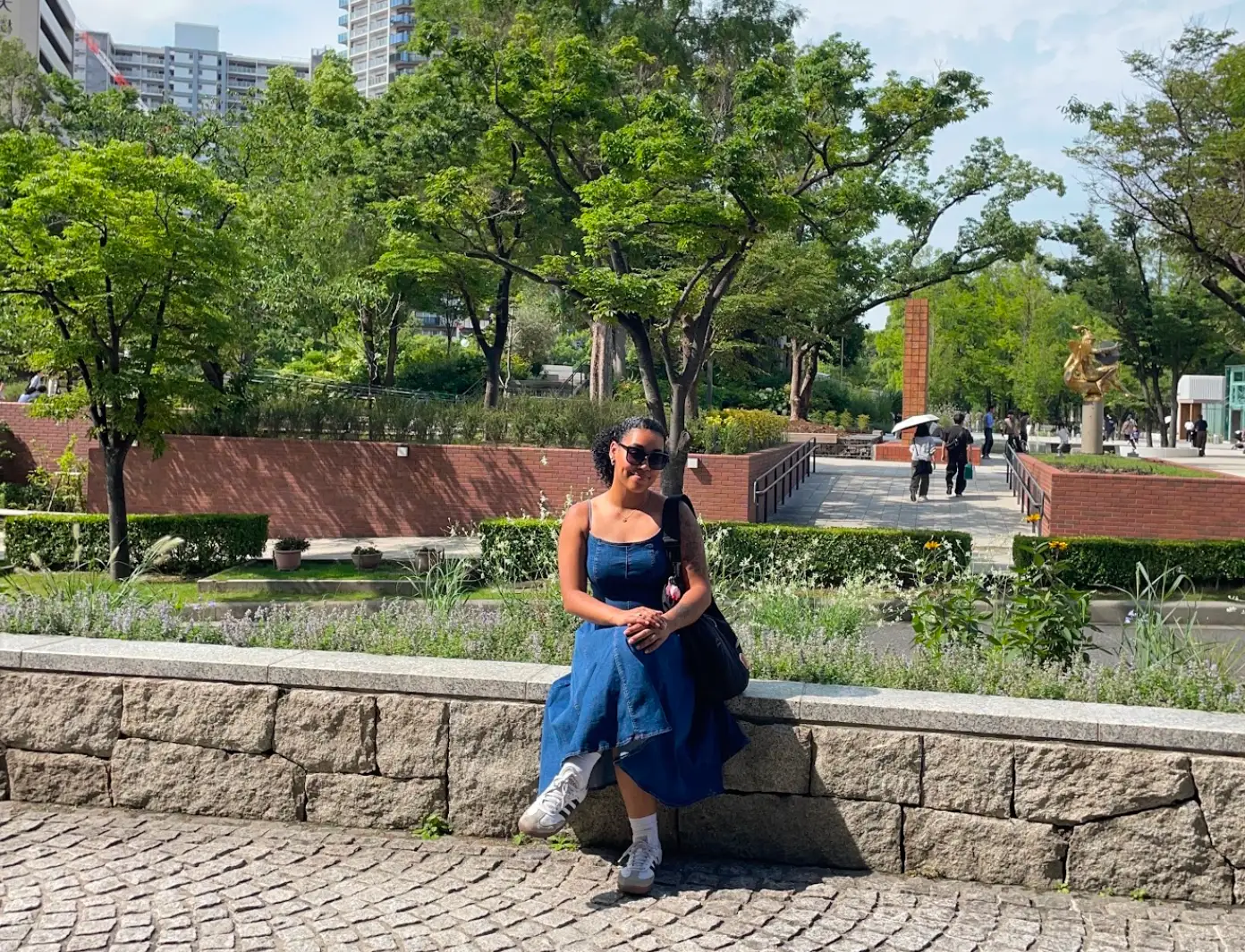 The writer wearing a blue dress and white sneakers and sitting on a ledge in Japan.