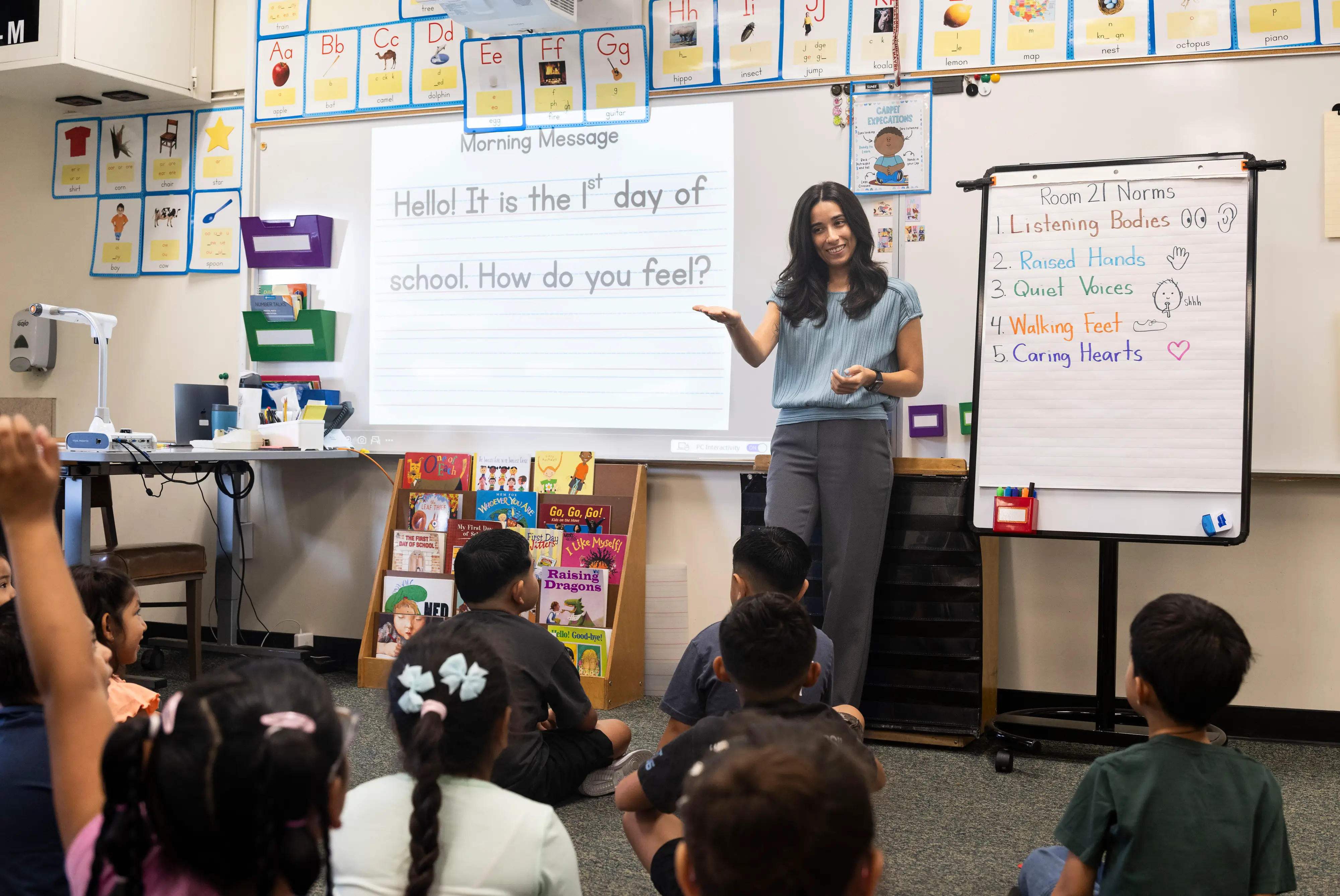 A woman in a blue shirt and gray pants stands before a sitting class of elementary students.