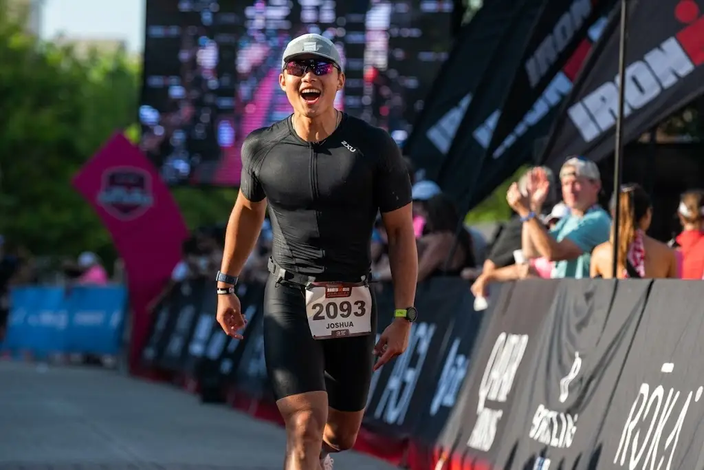 Triathlete wearing bib 2093 runs on an IRONMAN course past spectators and branded barriers.