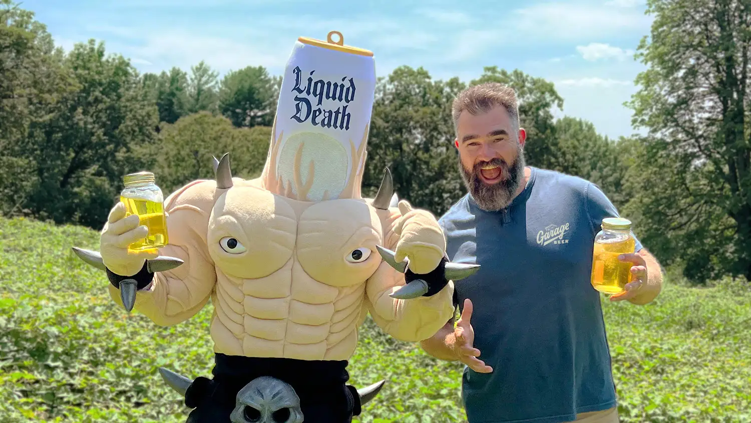 Jason Kelce poses in a field beside a Liquid Death can mascot while both hold jars of yellow liquid.