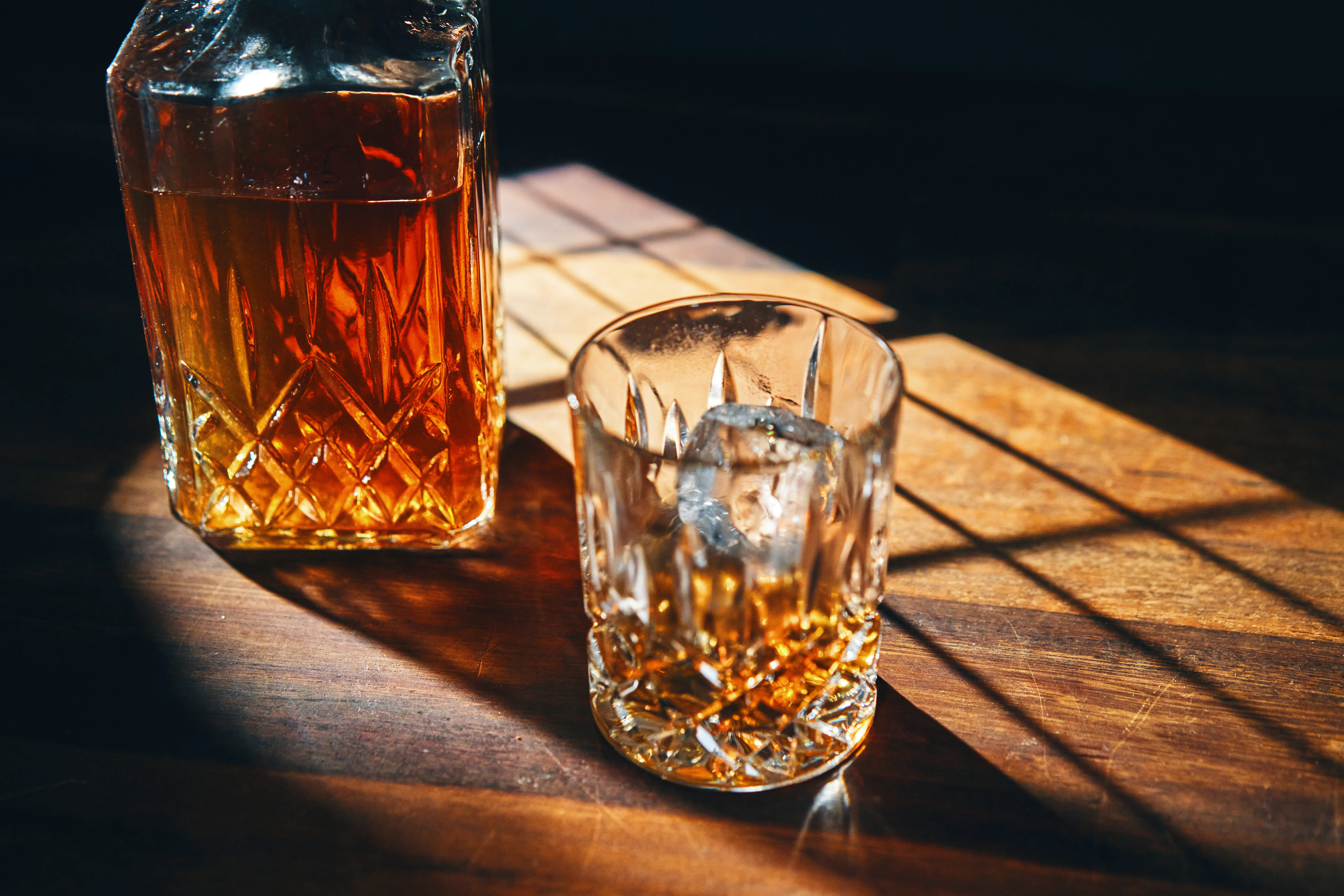 Cut-glass decanter and tumbler of amber liquor sit on a sunlit wooden table with dramatic shadows.