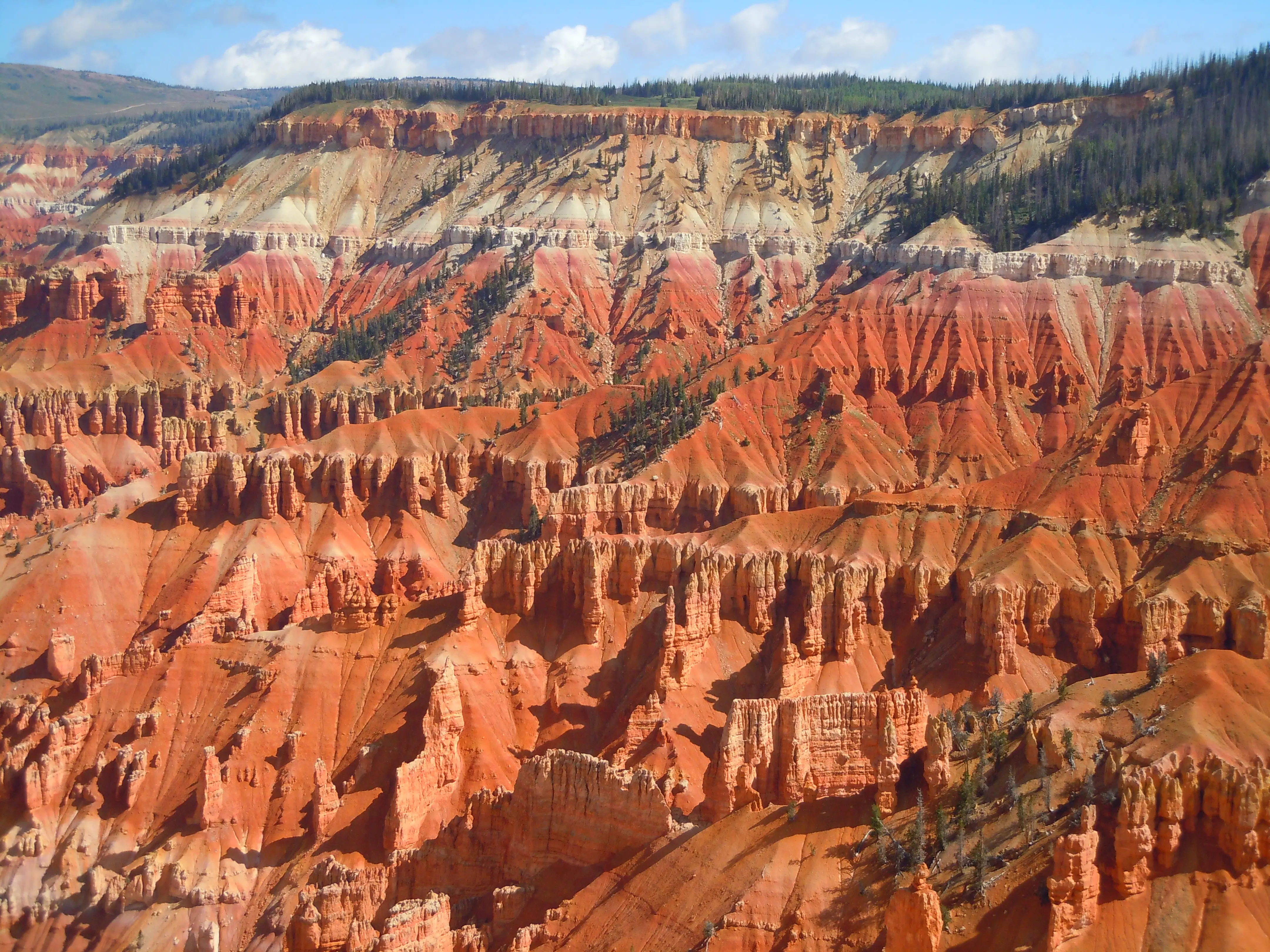 Layered red and tan rock formations at Cedar Breaks National Monument fill a rugged canyon beneath a forested rim and blue sky.