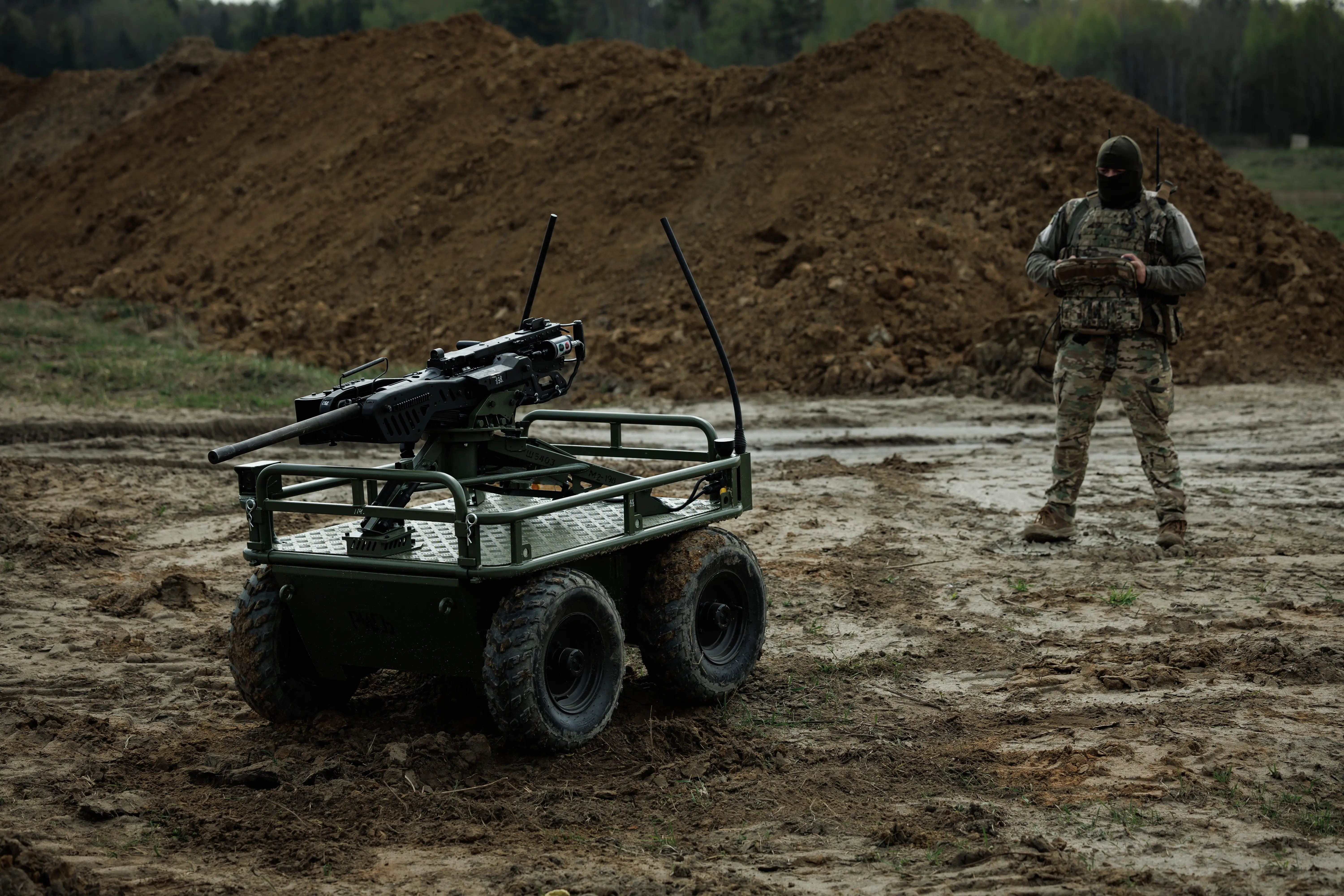 Camouflaged operator controls an armed unmanned ground vehicle on a muddy training field near large dirt mounds.
