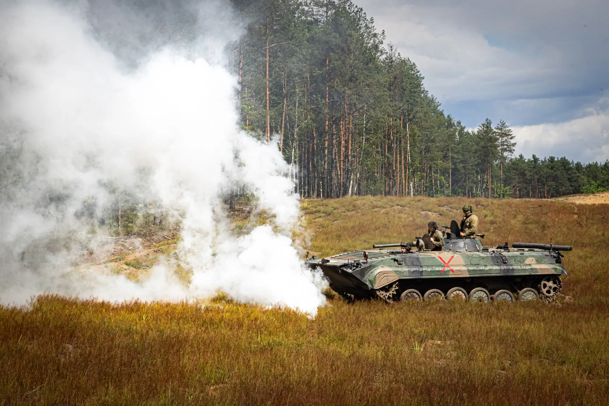 Tracked armored vehicle with uniformed personnel moves through a smoky grassy field beside a pine forest.