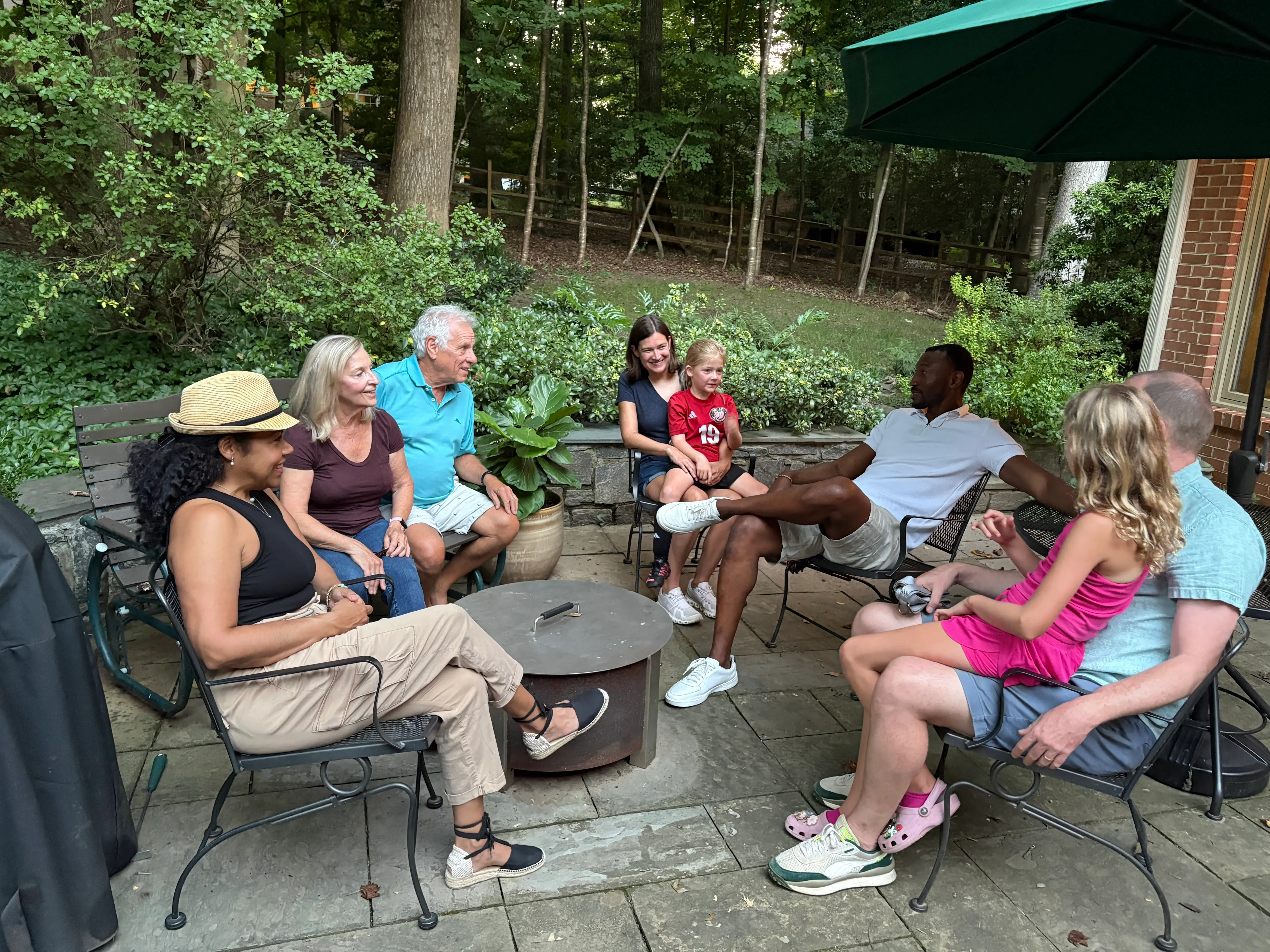 A group of people of all ages, including kids, seated on porch chairs in a backyard.