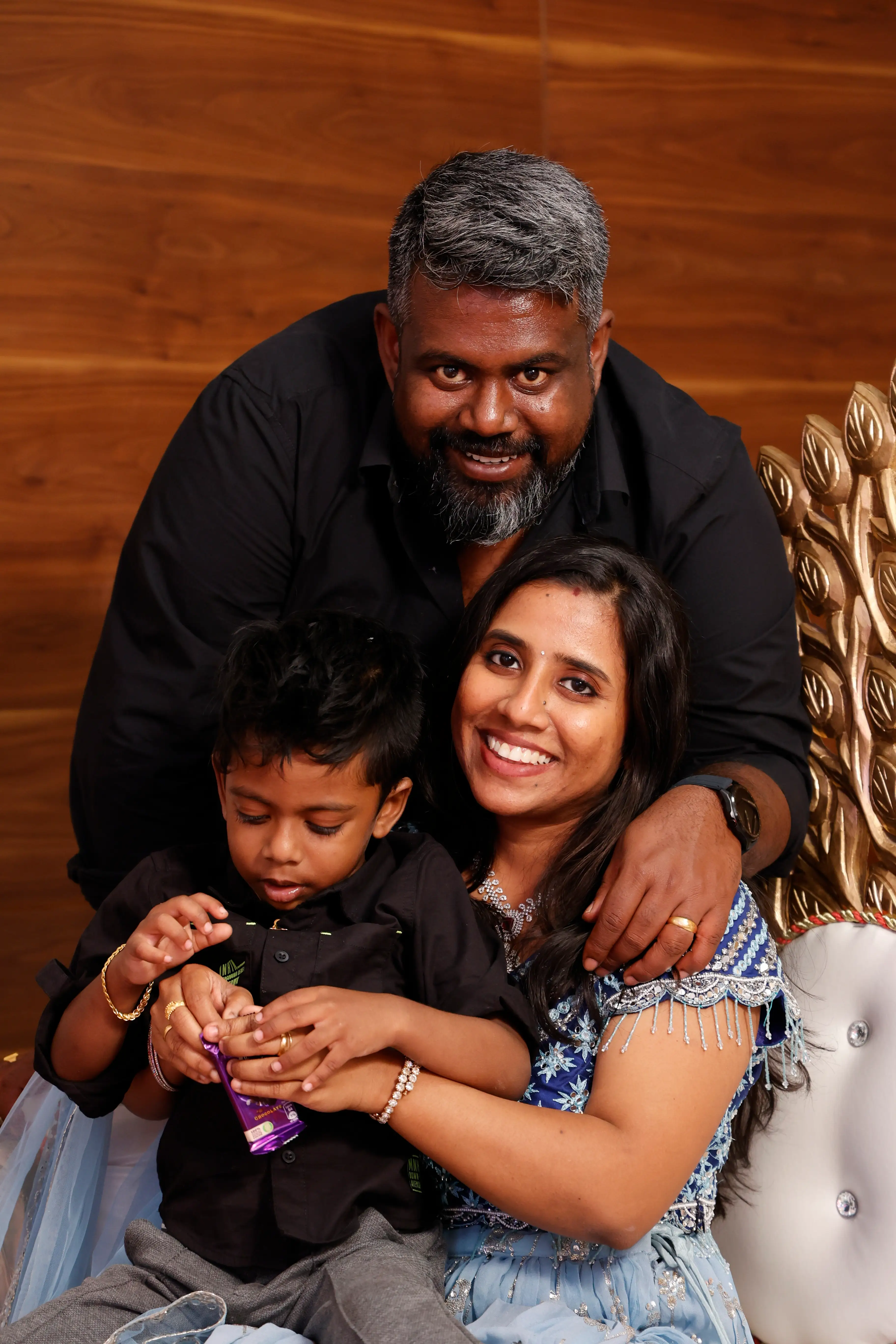 Mounika Chinnam is sitting down and holding her son. Her husband is standing and leaning on her shoulder.