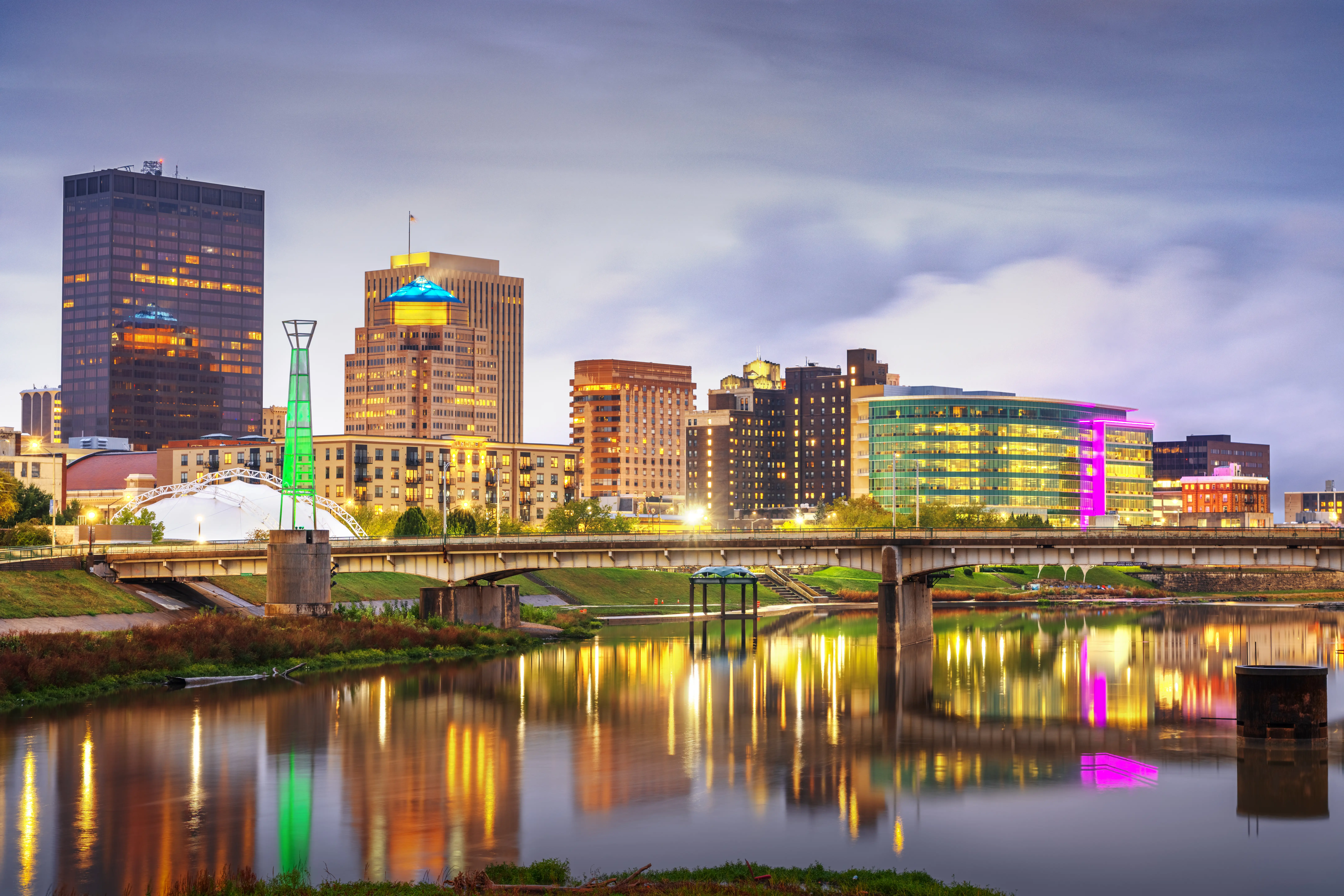 Downtown Dayton skyline glows at dusk beside the Great Miami River with bridges and colorful reflections.
