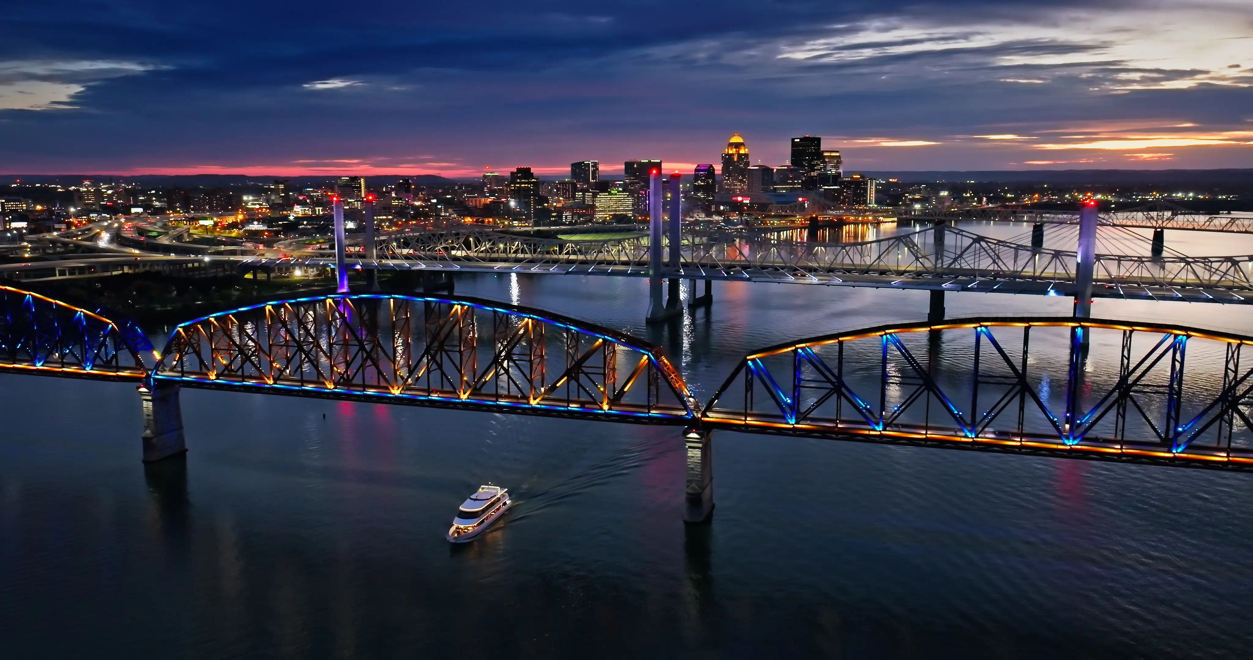 Downtown Louisville skyline glows at dusk behind illuminated bridges over the Ohio River with a boat below.