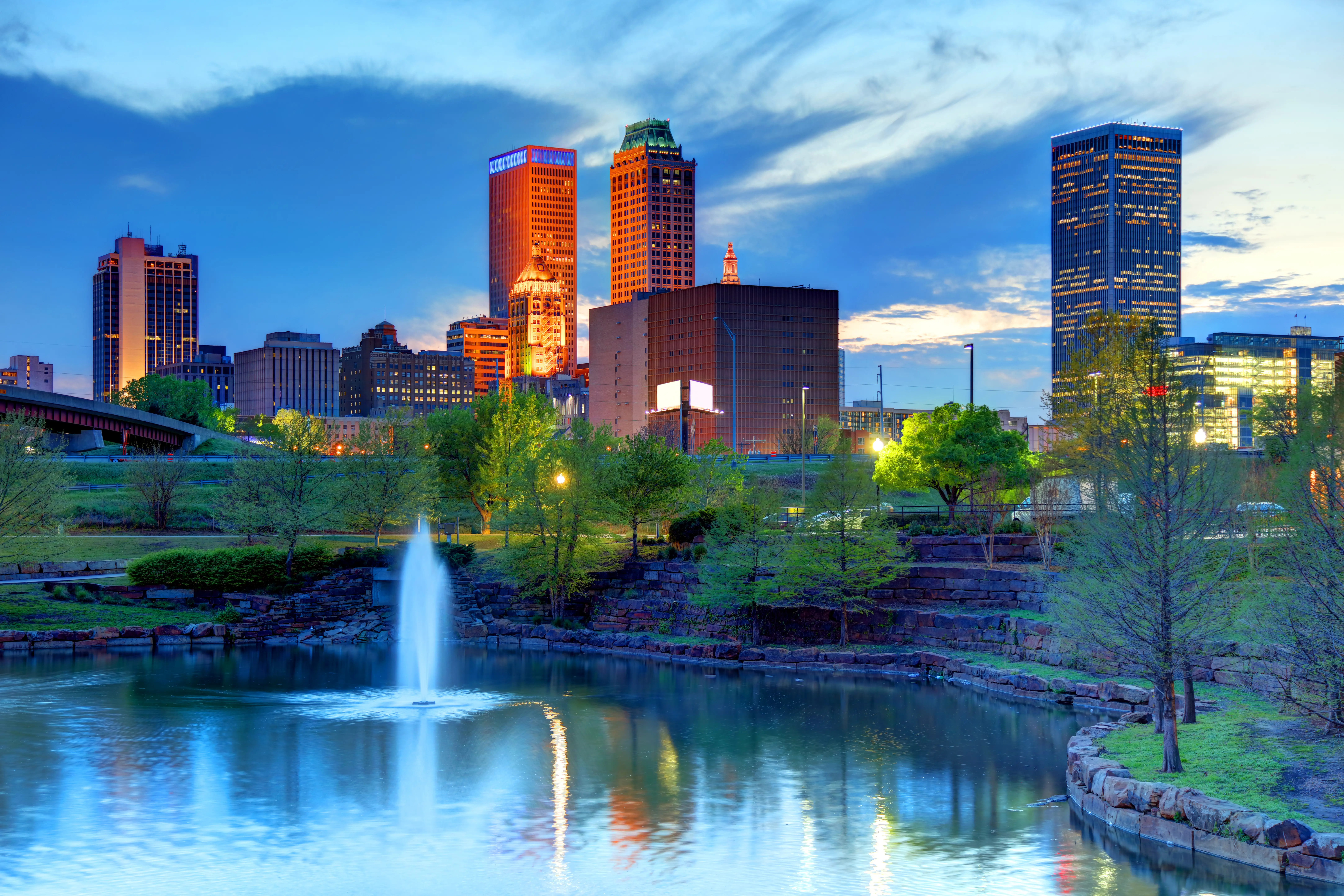 Downtown Tulsa skyline glows at dusk behind a park pond with a fountain and reflected city lights.