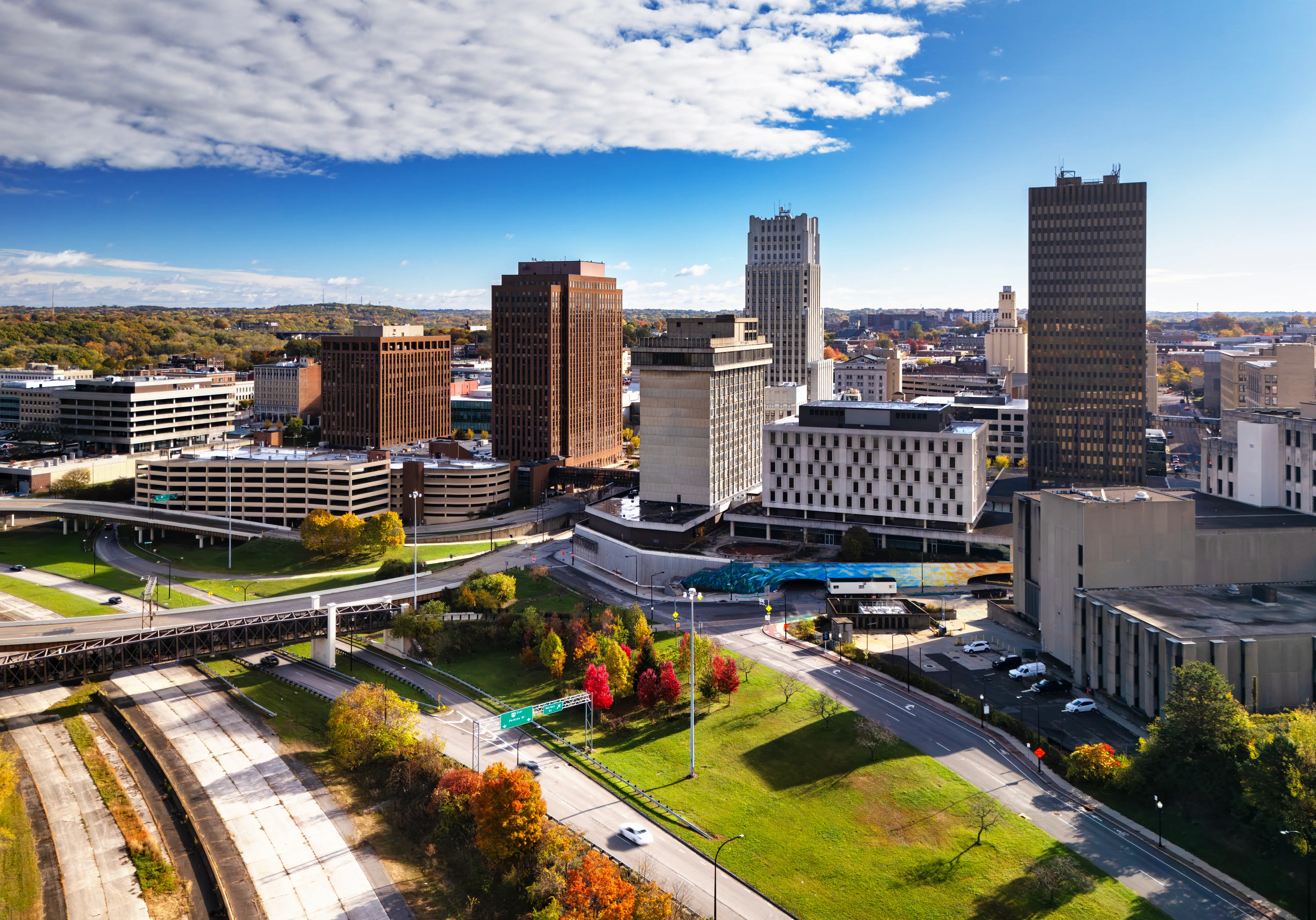 Downtown skyline with high-rise buildings, roads, green spaces, and autumn trees under a partly cloudy blue sky.