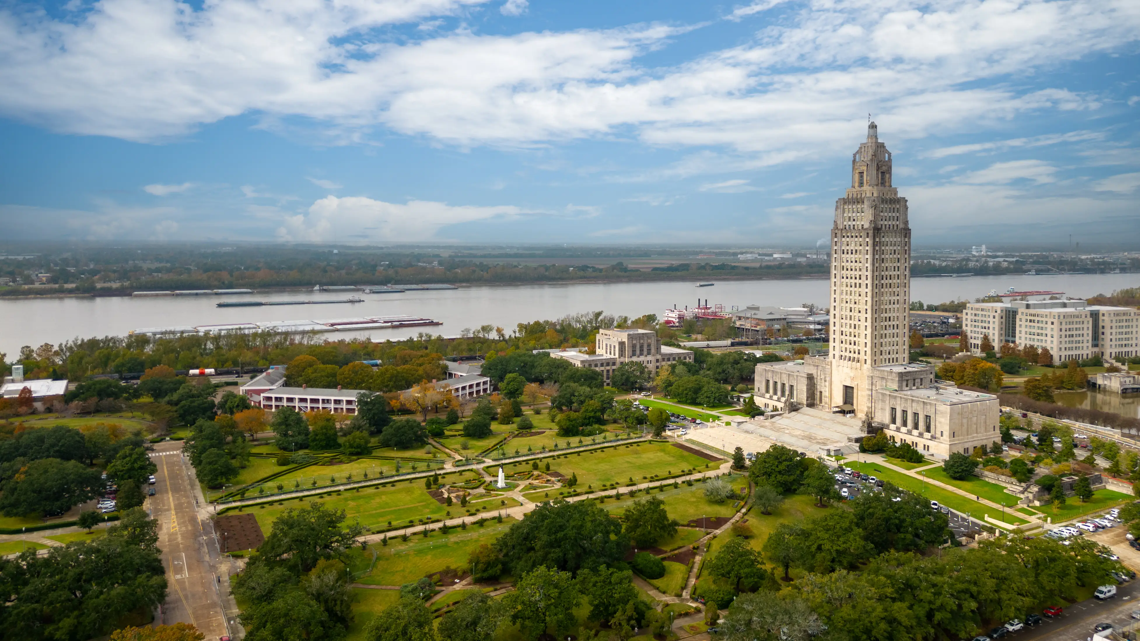Aerial view of the Louisiana State Capitol and surrounding gardens beside a broad river under a partly cloudy sky.