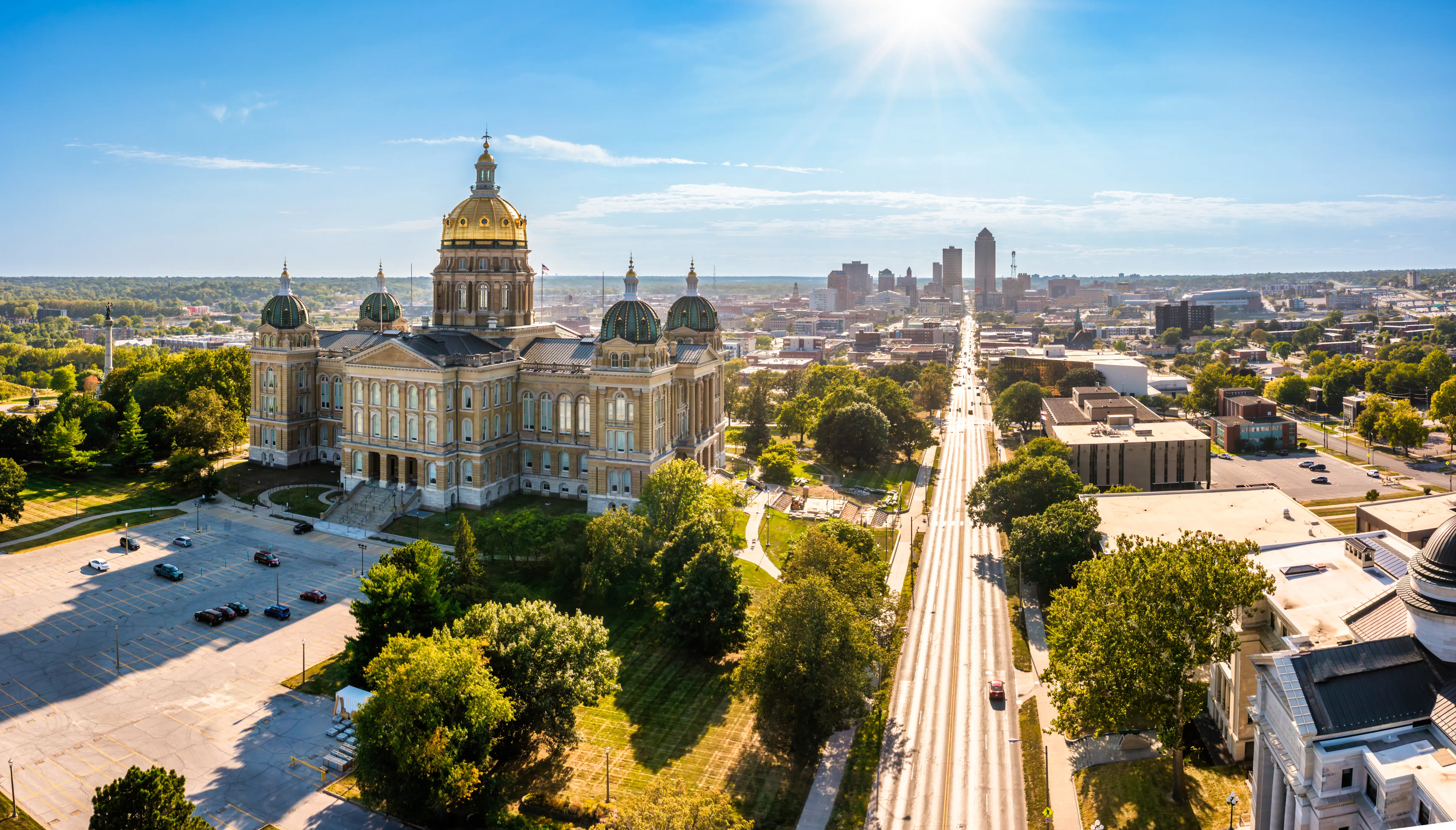 Iowa State Capitol with gold dome overlooks a broad avenue and downtown Des Moines under bright sun.