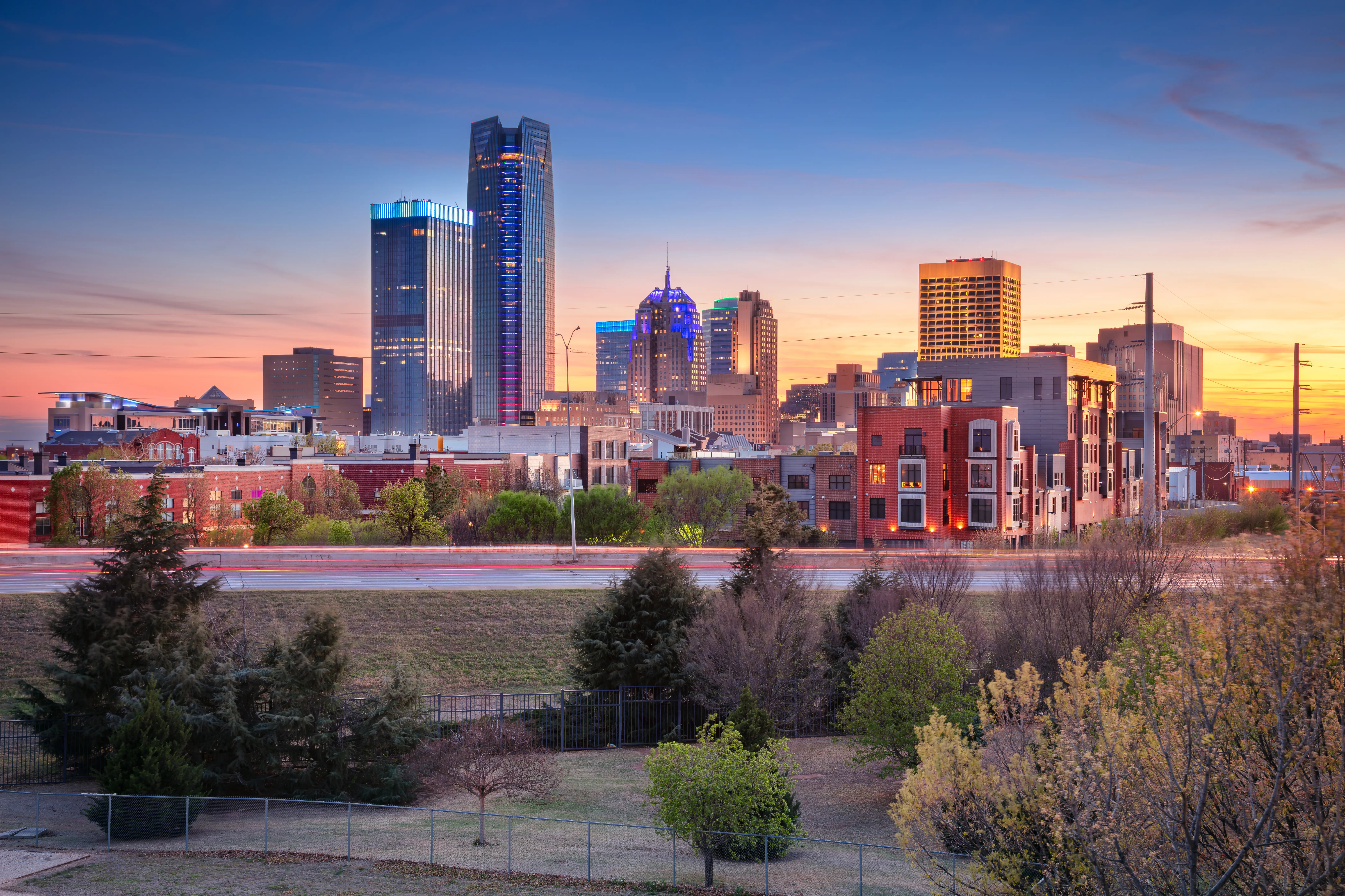 Downtown Oklahoma City skyline at sunset with high-rise towers, urban buildings, trees, and light trails in the foreground.