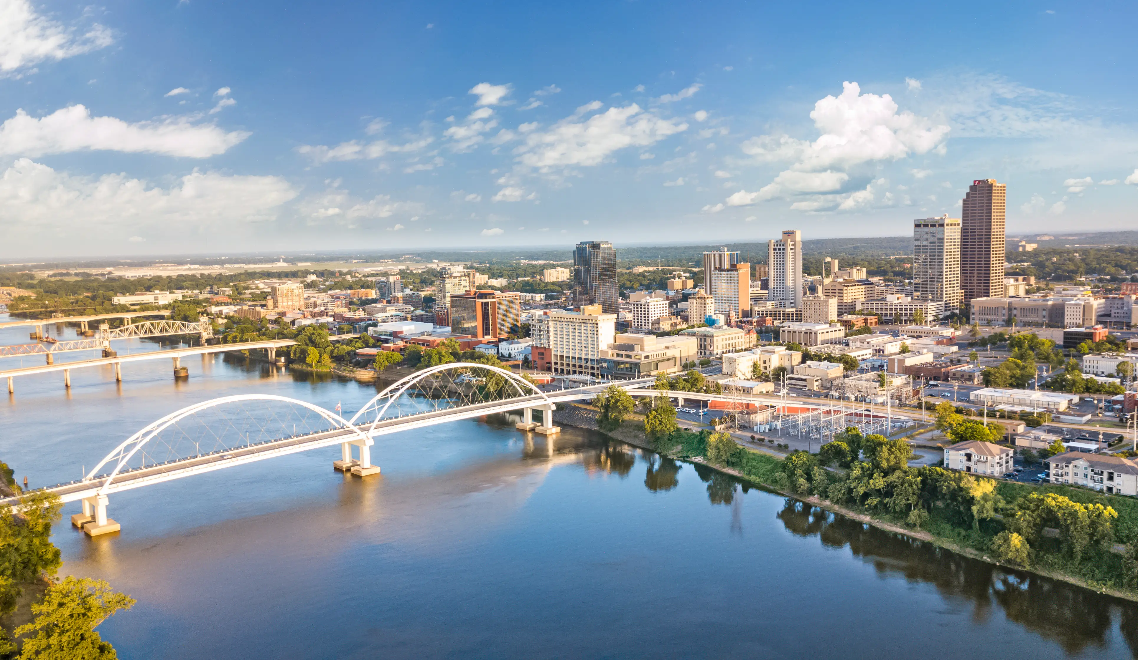 Downtown Little Rock skyline and a white arch bridge span the Arkansas River under a blue sky.