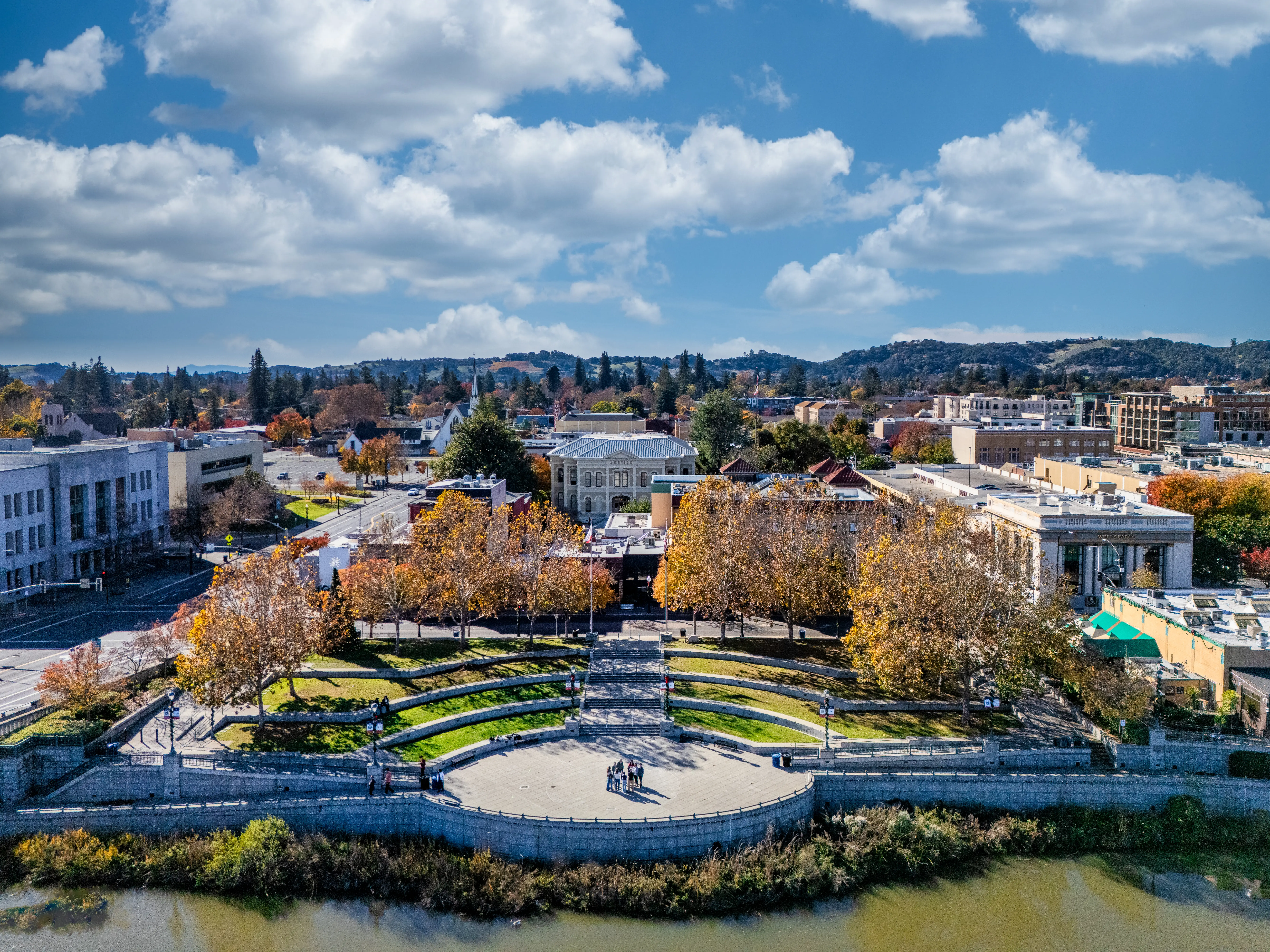 Aerial view of a landscaped riverside plaza with autumn trees, civic buildings, and cloudy blue sky.