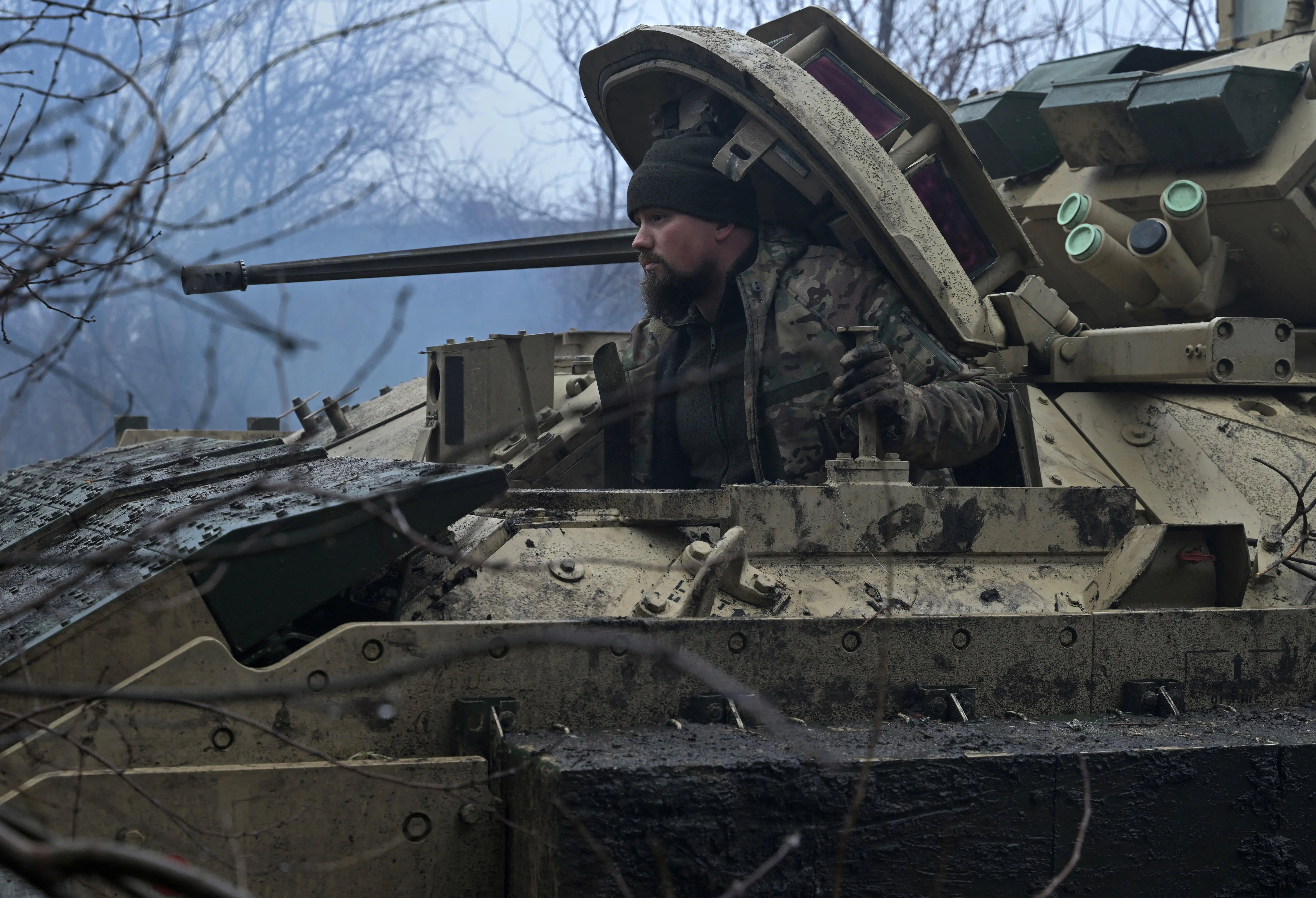 A helmeted person looks out from a muddy armored vehicle in a leafless wooded area.
