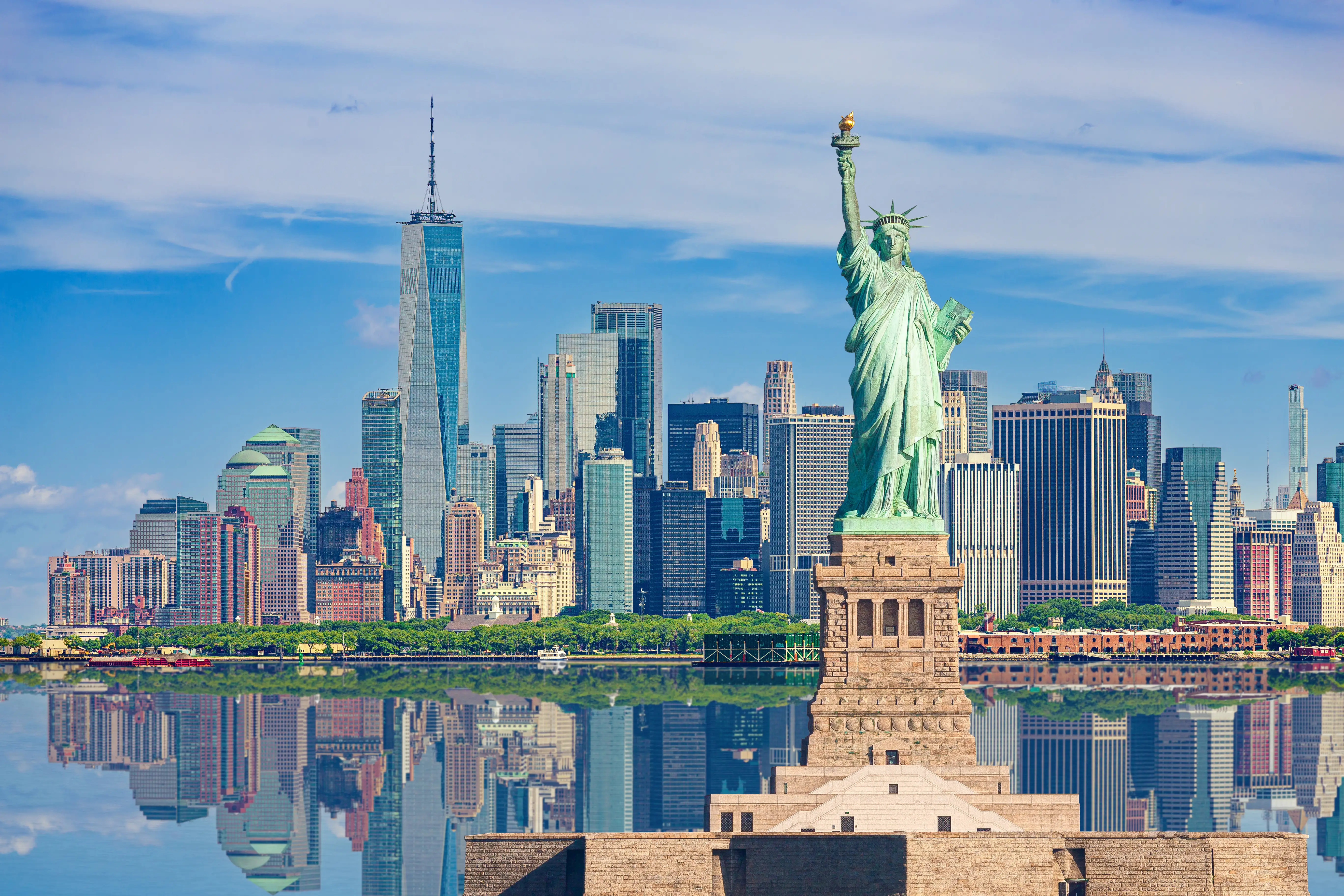 Statue of Liberty stands before the Lower Manhattan skyline reflected across calm water under a blue sky.