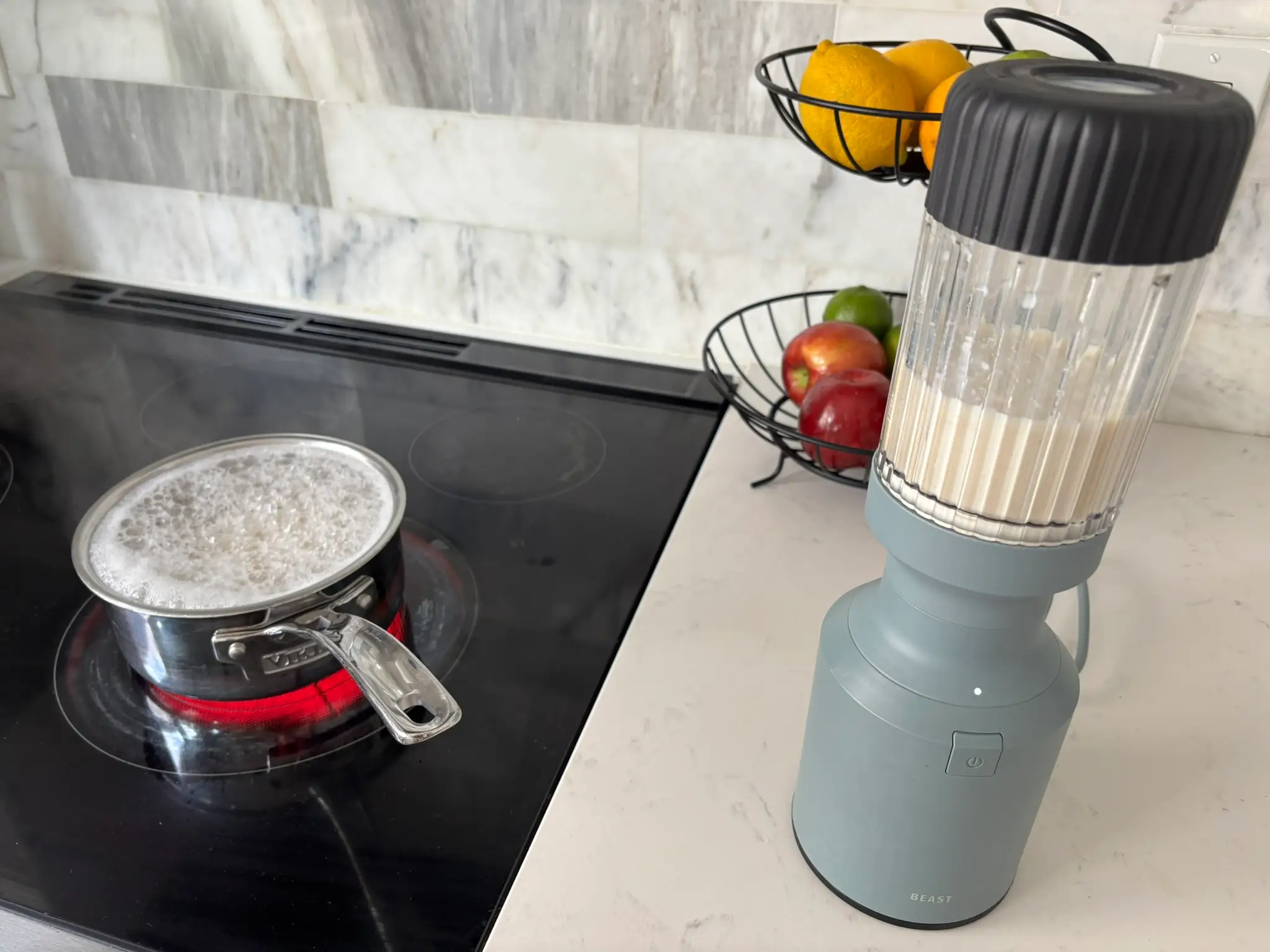 A pot of pasta boils on an electric cooktop beside a blender that is mixing a mac and cheese sauce.