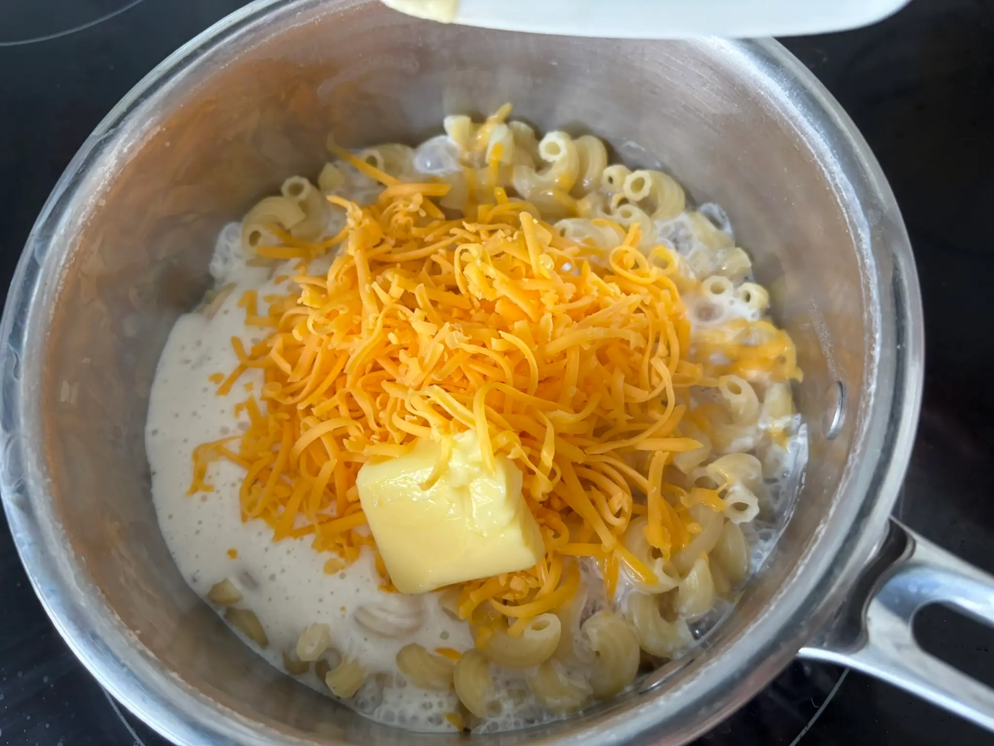 Elbow macaroni cooks in a saucepan with shredded cheddar, butter, and milk on a stovetop.