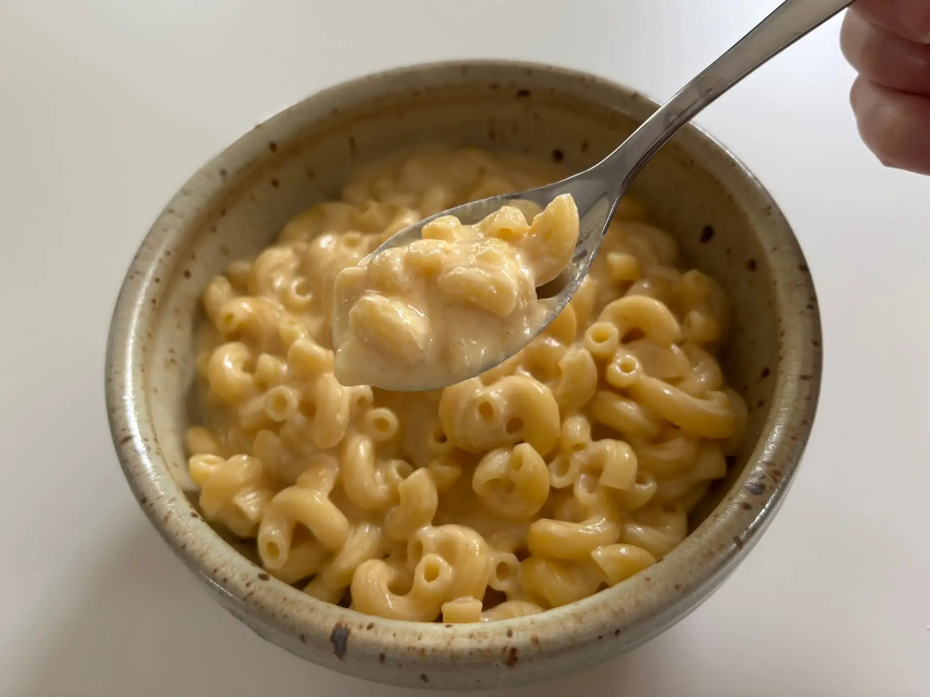 Spoonful of creamy macaroni and cheese held above a speckled ceramic bowl on a light tabletop.