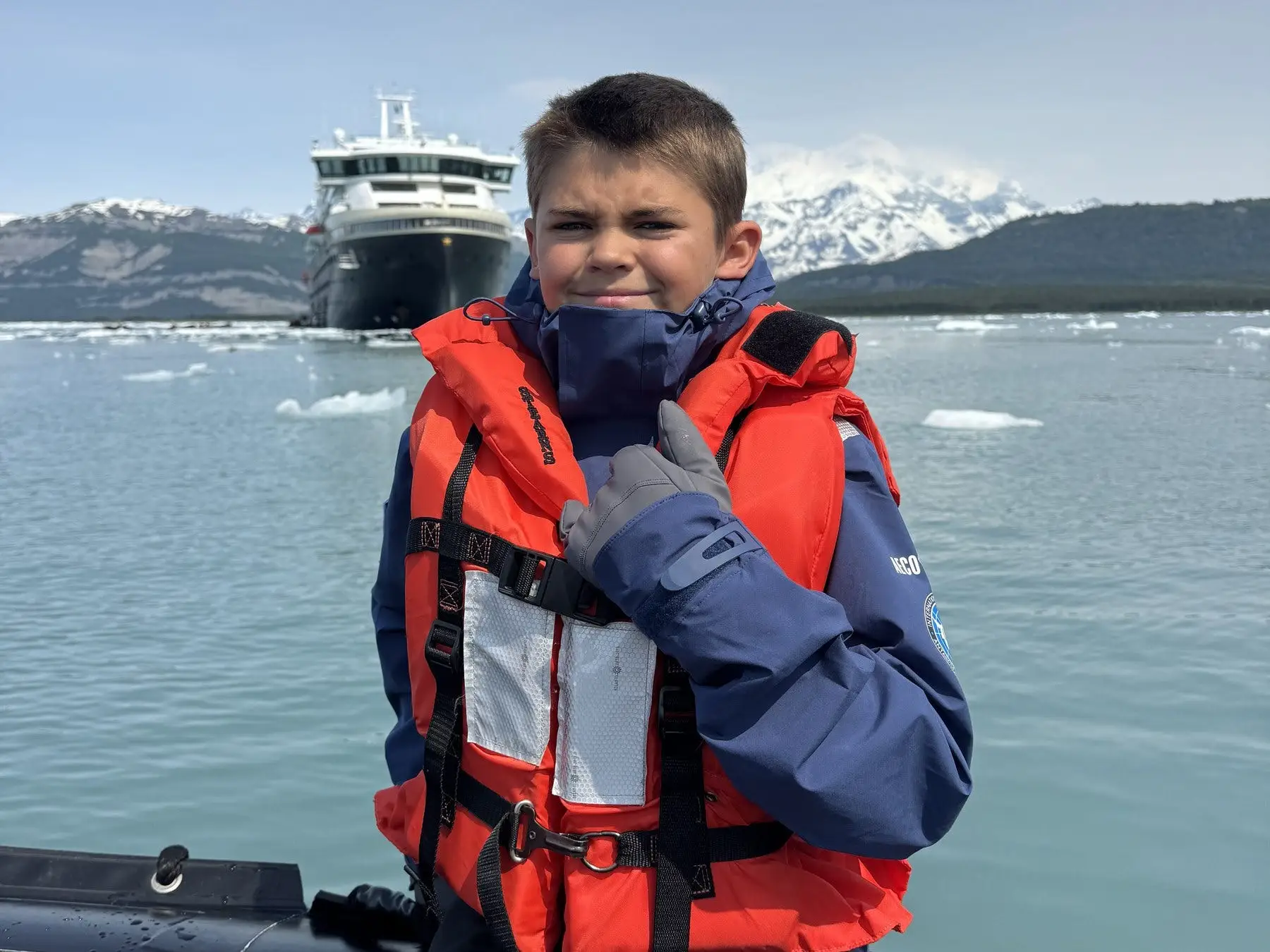 Kid in life jacket in Alaska, smiling with ship in distance