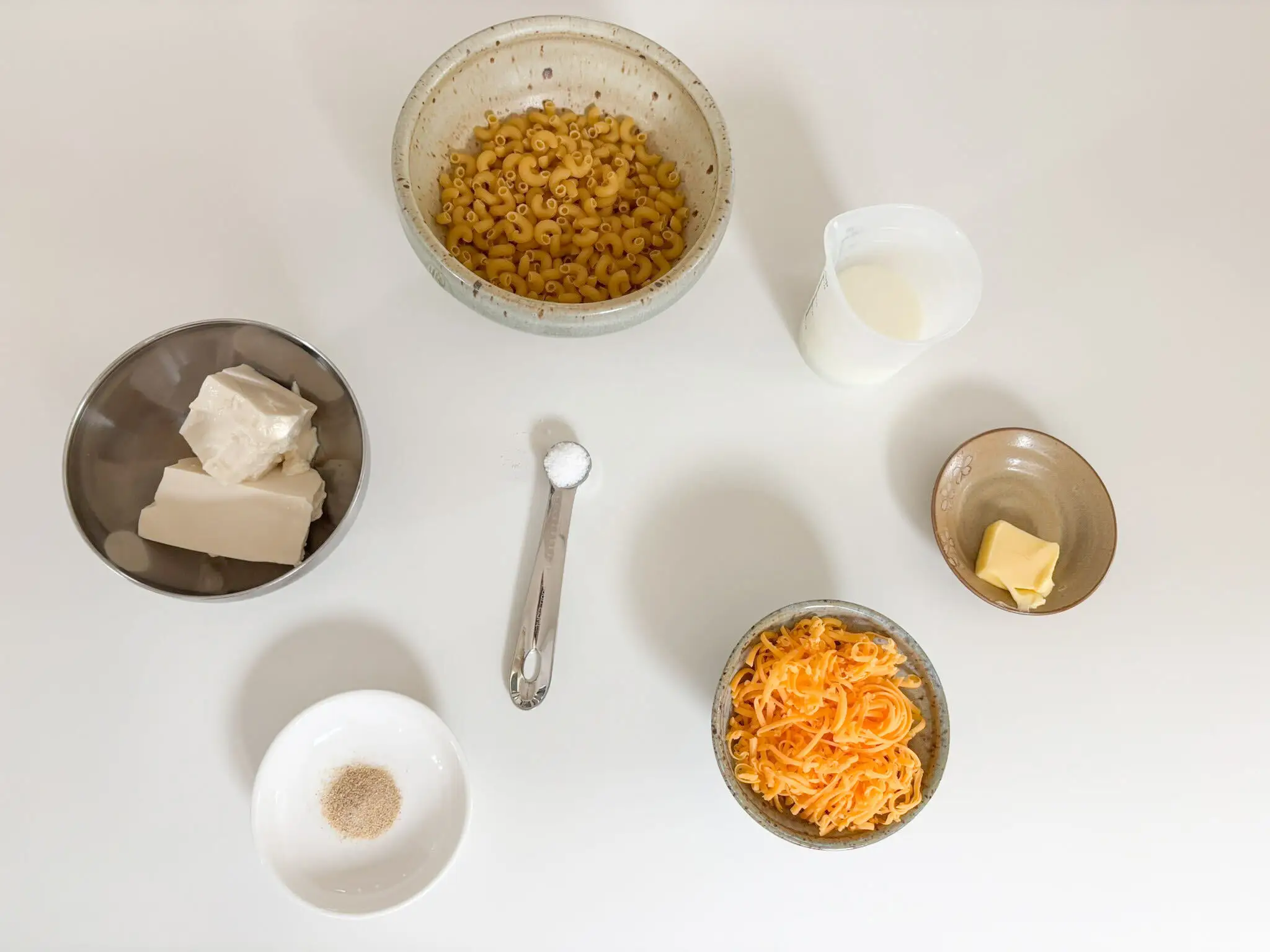 Overhead arrangement of mac and cheese ingredients, including shredded cheese, milk, butter, salt, garlic salt, and tofu.